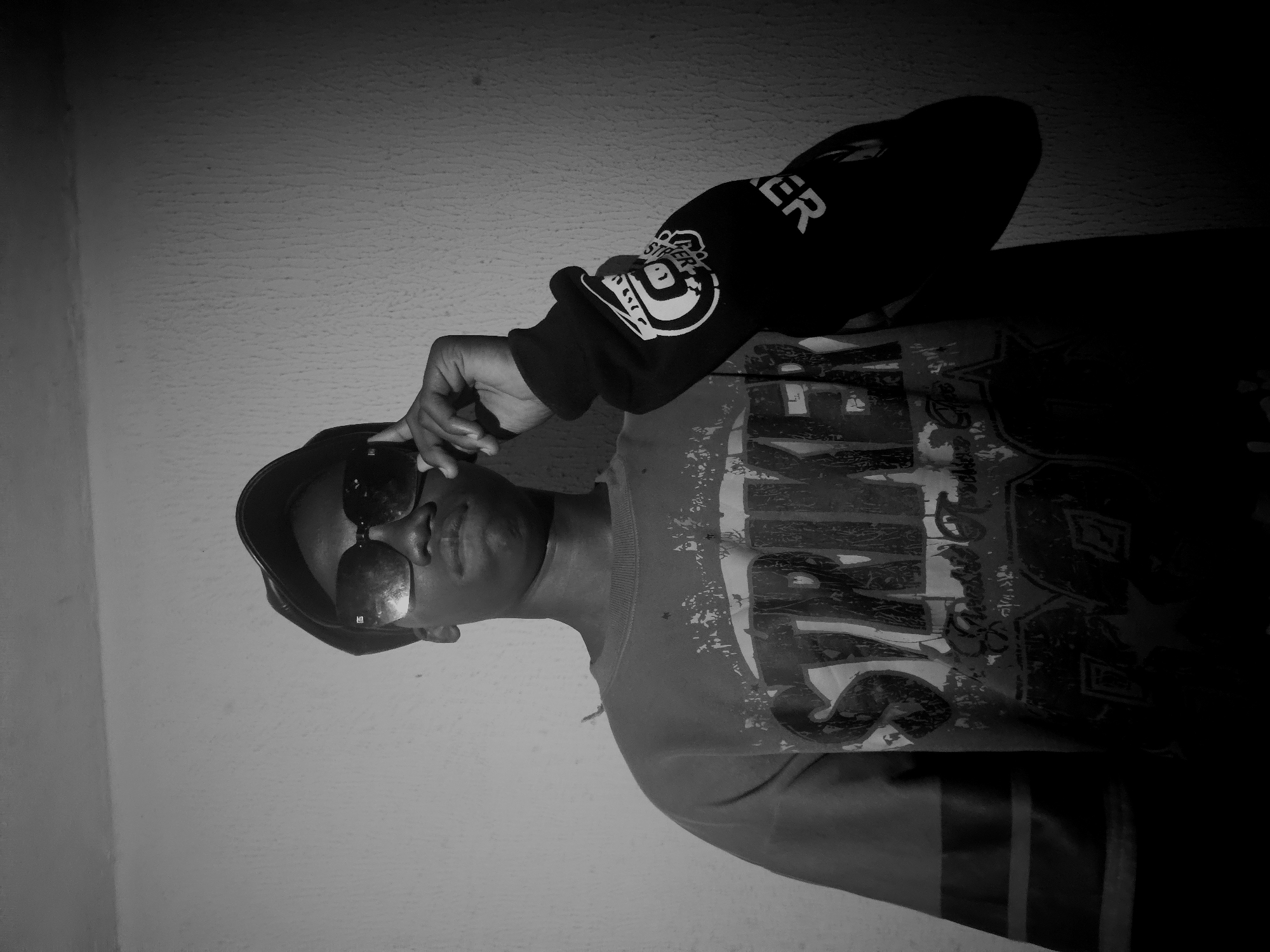The image depicts a person wearing a black cap and sunglasses, positioned indoors against a pale wall. The individual is wearing a t-shirt with a large, stylized graphic that includes the word "STRIKERS," with a black long-sleeved hoodie that has a graphic on the sleeve. The person is holding the sunglasses with their hand, suggesting they are adjusting them. The image has been rotated, so the person appears to be lying horizontally. The background is plain, focusing the attention on the individual and their attire. There is no specific evidence that places the image in Nkwelle, Nigeria.