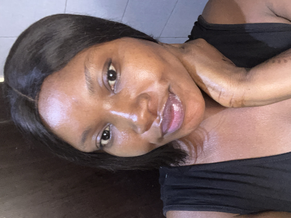 A close-up, chest-up portrait shows a female individual in Dzhidu, Nigeria, against an indistinct indoor background. The subject has dark skin, dark, straight hair parted in the center and reaching approximately shoulder length. She is wearing a dark, sleeveless garment. Her face exhibits a glossy appearance, particularly on the forehead, nose, and cheekbones. Her lips are full and coated with a light pink or purplish gloss. Her dark eyes are open and directed towards the viewer. The individual's right hand is visible, with fingers resting against her neck and lower jawline. The background consists of a dark, uniform surface on the left and a lighter, potentially tiled or painted wall on the right.