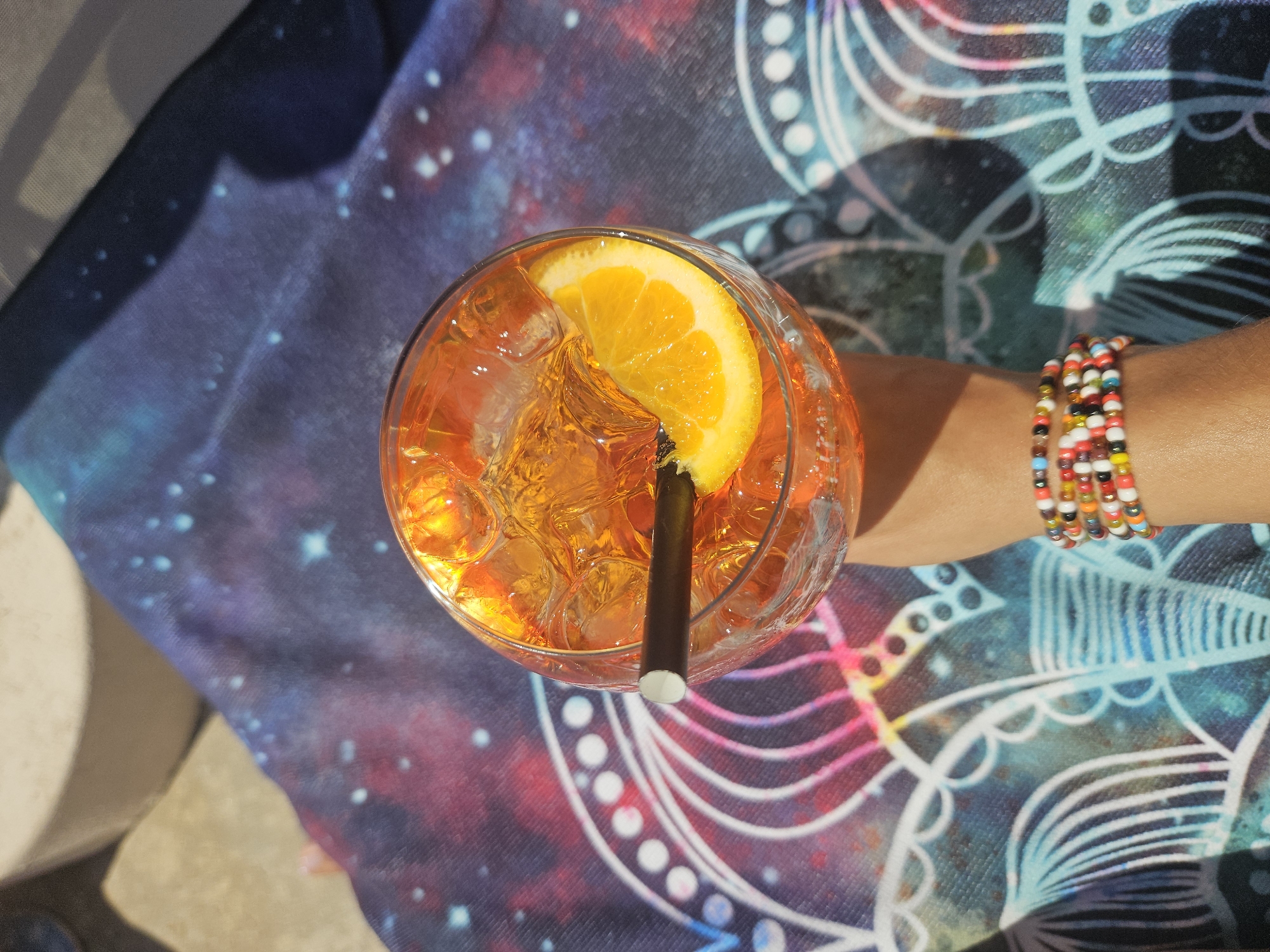 A close-up overhead shot shows a hand holding a glass of orange-colored drink filled with ice and a slice of orange. A straw is visible in the drink. The glass rests on a fabric with a galaxy-like pattern and abstract designs. The hand is adorned with a colorful beaded bracelet. The lighting suggests it is daytime, with shadows indicating sunlight. The background is out of focus, showing a hint of a textured surface.