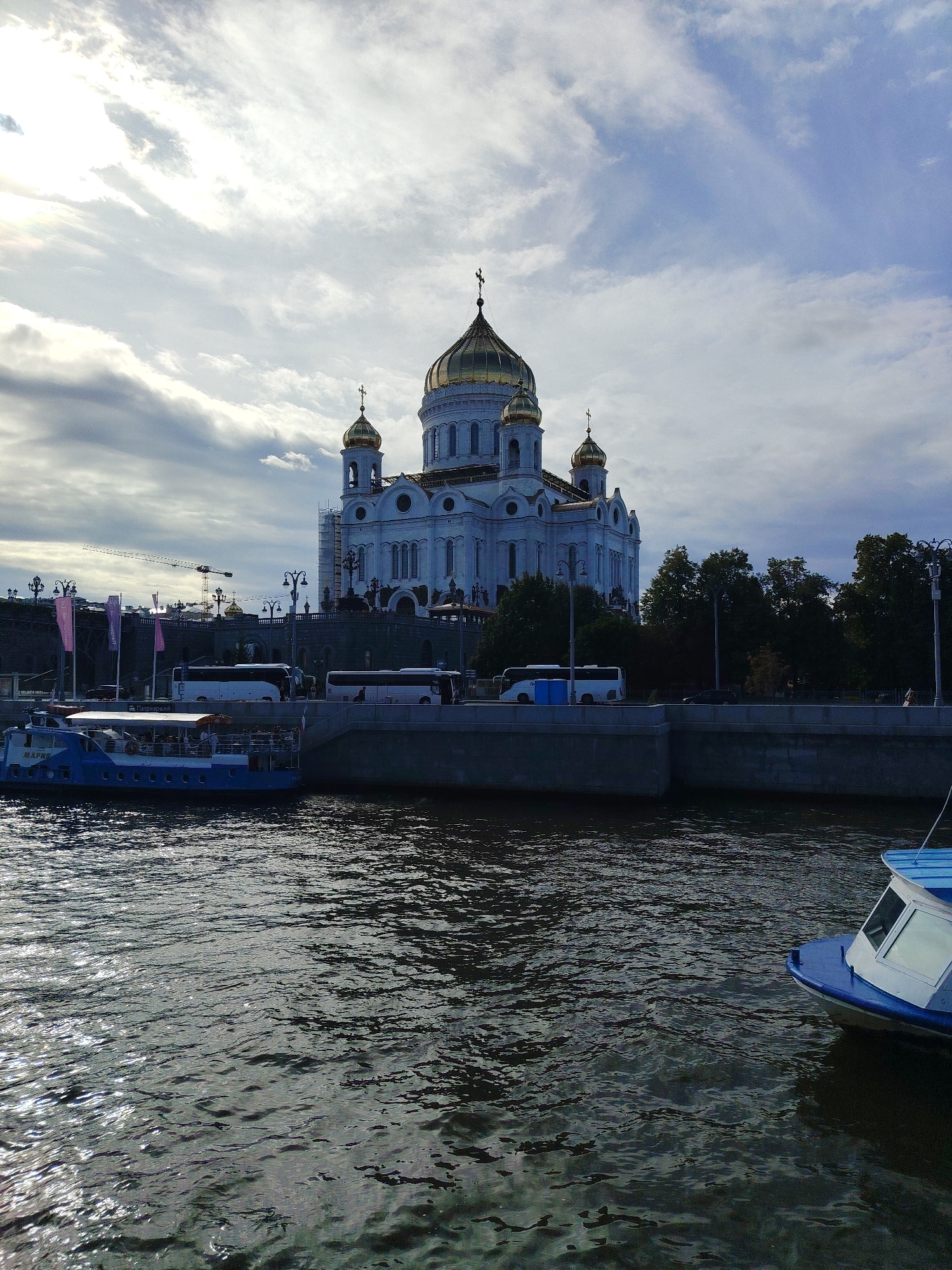 The image depicts a wide view of the Moskva River in Moscow, Russia, with the Christ the Saviour Cathedral prominently featured on the embankment. The cathedral, a grand white structure with golden domes, is the focal point, its architectural details clearly visible under a cloudy sky. Several white tour buses are parked along the embankment in front of the cathedral, suggesting a popular tourist area.
In the foreground, the river is choppy with sunlight glinting off the ripples. Two boats are visible on the water. A larger blue and white passenger boat, possibly a tour boat, is docked near the embankment on the left side of the image. A smaller white and blue boat is partially visible on the right side of the frame, closer to the viewer.
The embankment itself is lined with trees, lampposts, and a stone wall. Construction cranes are visible in the background behind the cathedral, indicating ongoing development or restoration work. The sky is a mixture of bright clouds and patches of blue, suggesting it could be late afternoon or early morning. The overall scene conveys a sense of historical grandeur combined with modern city life.