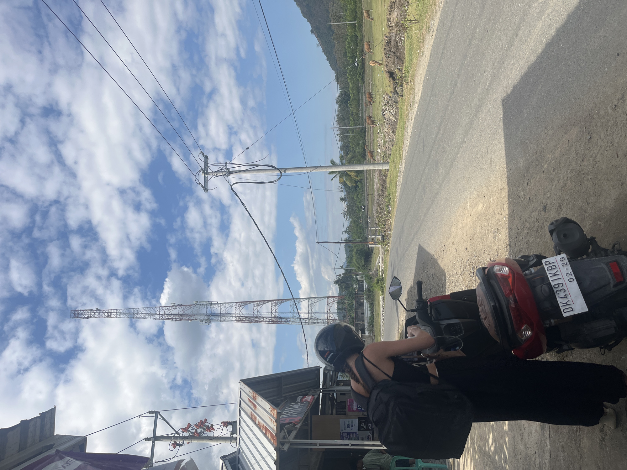 A person wearing a black top, black pants, a black helmet, and a black backpack stands next to a red and black motorcycle on a dirt and asphalt road in Desa Lamunga, Indonesia. The person is looking down at their hands, which are holding a phone. The motorcycle has a license plate that reads "DK 4391 KBP 02-29". To the left of the road is a small building with a corrugated metal roof and a sign that reads "TRIMAKASI". There are power lines and a tall cell tower in the background. A herd of brown cows grazes in a field to the right of the road, and a lush green hillside with trees is visible beyond the field. The sky is blue with scattered white clouds.