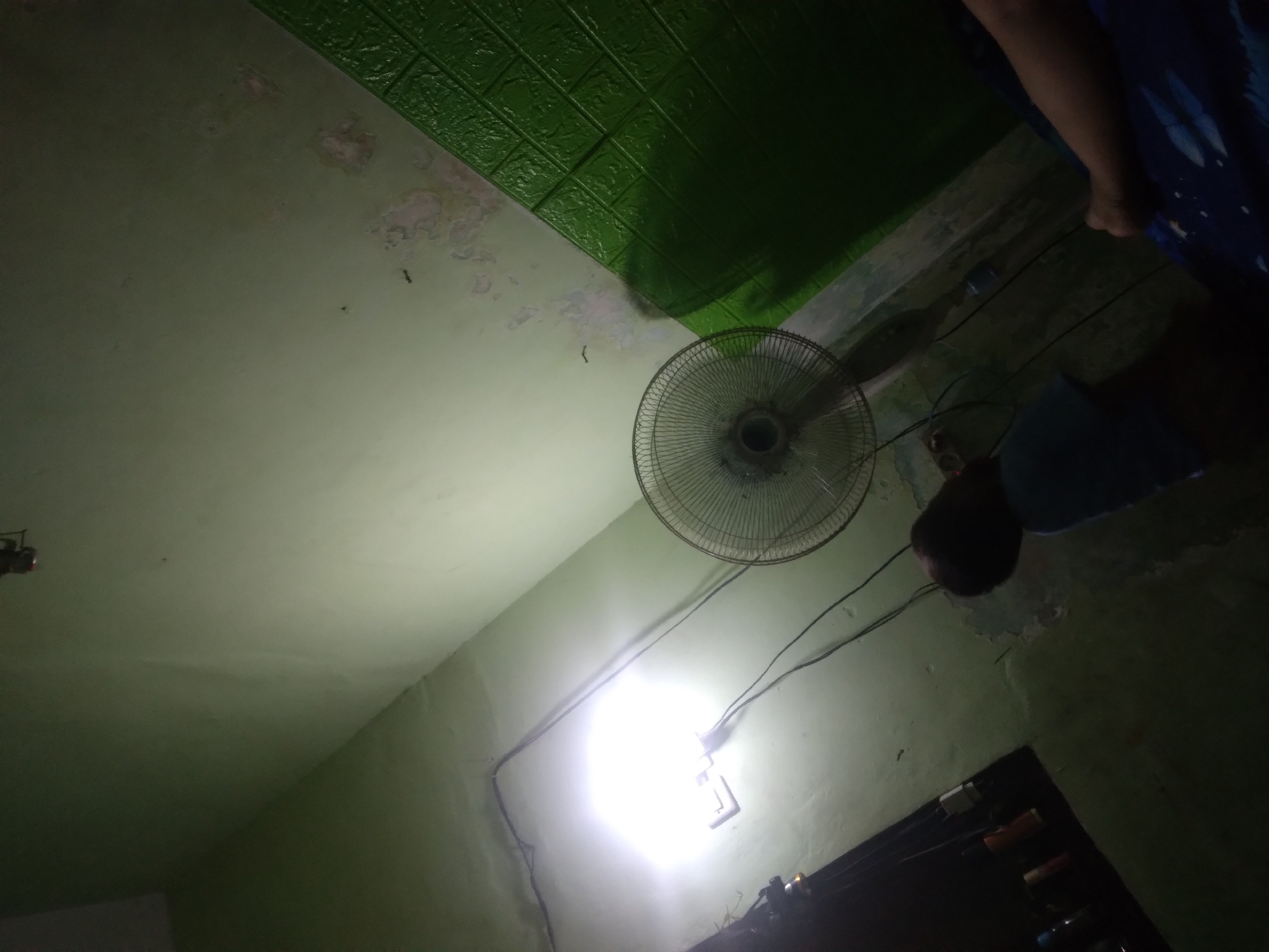 The image shows an indoor scene, likely a room, with a light green wall. A bright light source, possibly a lamp, is illuminating a portion of the wall and a white electrical outlet. A ceiling fan is visible on the right side of the frame. A dark-colored fan with a wire grille is positioned in the center. The wall has peeling paint in some areas. To the right, a section of the wall is covered with green textured wallpaper with a brick-like pattern. In the lower part of the image, a person's head and shoulders are visible, turned away from the camera. Their body is partially obscured by shadow. On the far right, a person's arm is resting on a dark blue surface with a white pattern. The overall lighting is dim, with the exception of the bright light source. The scene is likely within a residential setting in Wringinanom, Indonesia, given the context provided.