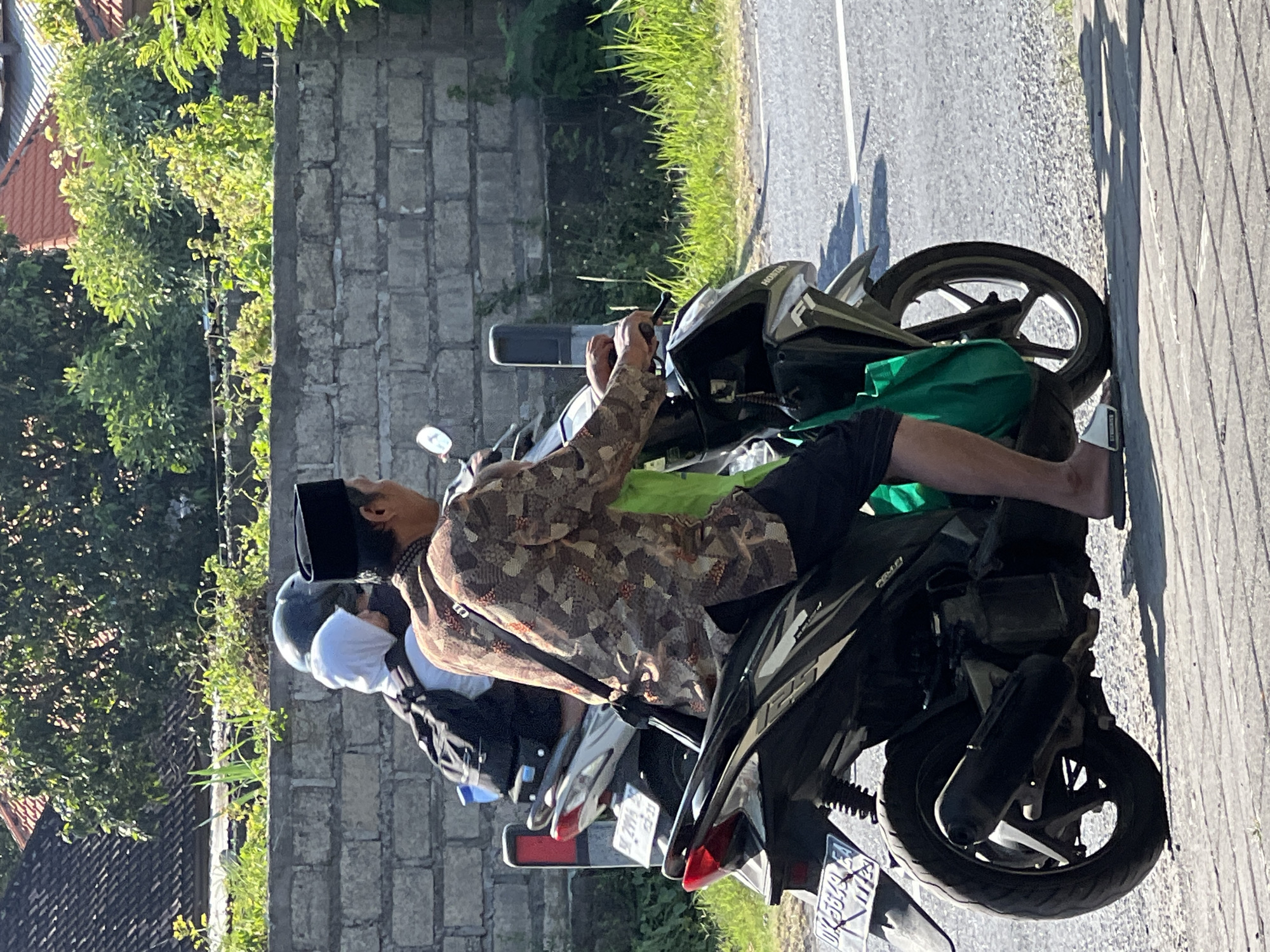 The image shows two people on a motorcycle driving on a paved road. The person in the foreground is a man wearing a batik shirt, black shorts, and a black peci hat. He is operating the motorcycle. Behind him, a second person, likely a woman, is wearing a white hijab and a dark backpack. They are traveling past a low, grey brick wall with green vegetation growing on top and behind it. In the background, residential buildings with terracotta roofs are visible. The road surface is asphalt mixed with gravel, and sunlight casts shadows, suggesting it is daytime. The license plate of the motorcycle appears to be from Denpasar, Indonesia, indicated by the "DK" prefix.