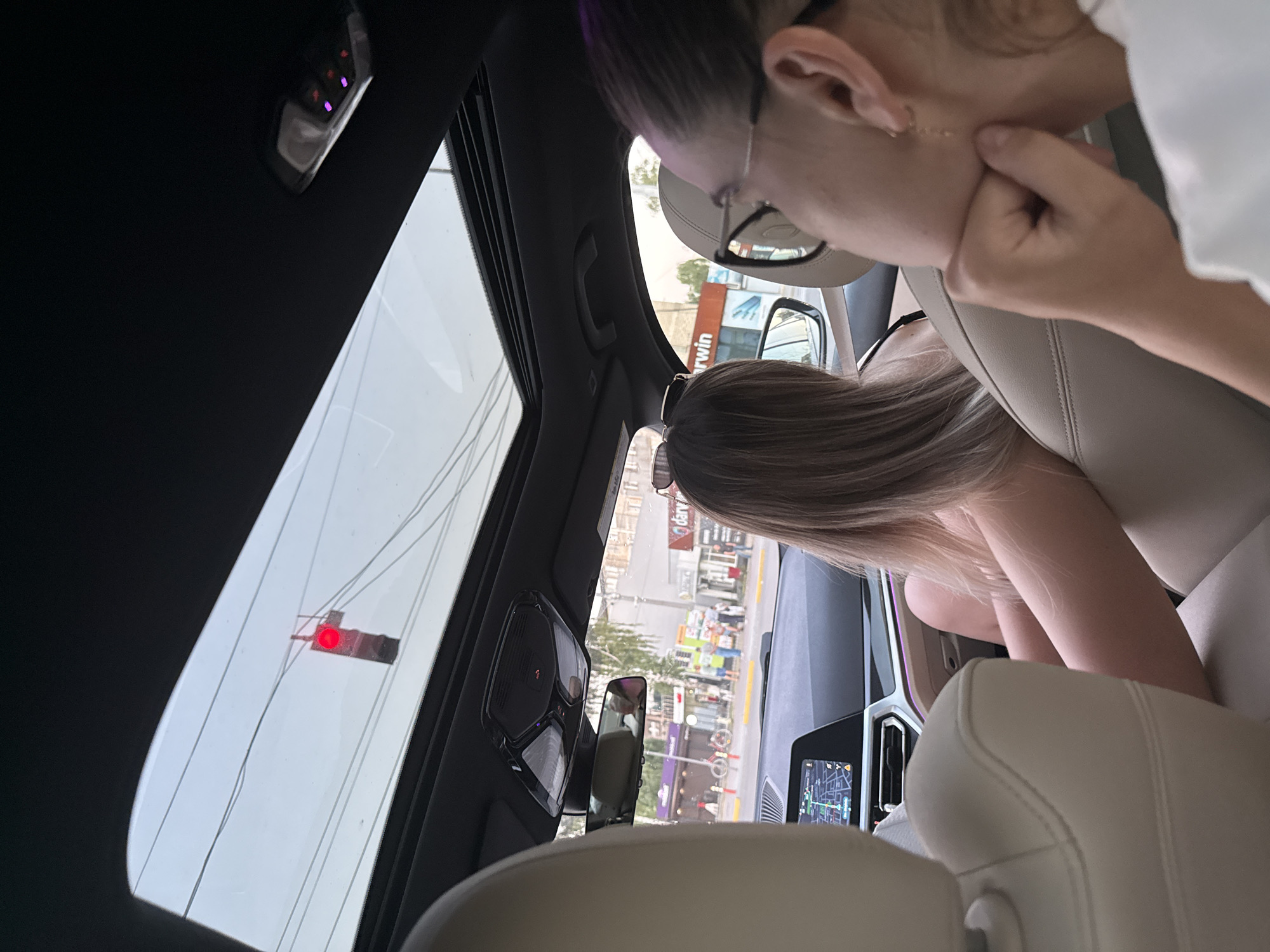 The image captures an interior view of a car driving through the city of Fundul Galbenei, Moldova. Through the car's sunroof and windshield, a red traffic light is visible, indicating a stop. Inside the car, two individuals are present. In the foreground, the side profile of a person wearing glasses is visible, resting their chin on their hand. In the middle ground, another person with blonde hair is seated, facing away from the camera. The car's dashboard displays a navigation screen. The external environment appears to be a street scene with buildings, trees, and pedestrians, suggesting an urban setting. The time of day seems to be late afternoon or early evening, given the lighting conditions.
