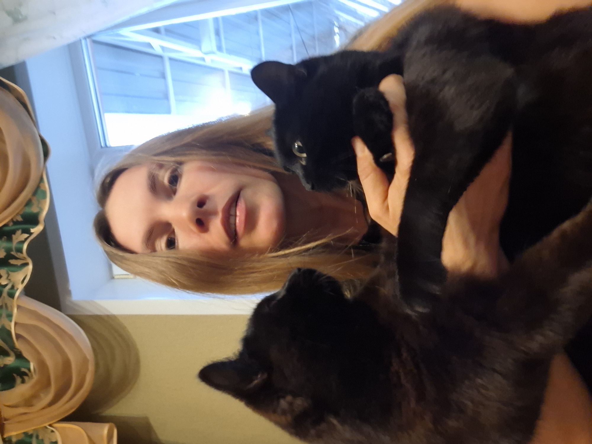 A woman with long blonde hair holds two black cats in her arms. The woman is fair-skinned, with brown eyes and a slight smile. The cats appear to be interacting with her and each other. A window is visible in the background, providing natural light. There's a patterned fabric draped to the side. The location, Khlevnoye, Russia is not definitively ascertainable from the visual information.