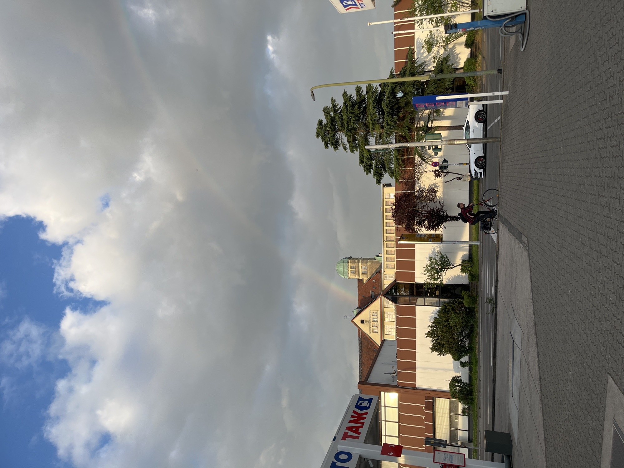 The image captures a street scene in Landau in der Pfalz, Germany under a cloudy sky with a partial rainbow visible. A person on a bicycle is positioned at a crosswalk near a gas station identified by a red and white "TANK" sign. Adjacent to the gas station is a multi-story building with a domed tower. A white car is parked further down the street, near a green streetlamp. The ground is paved with interlocking stones.
