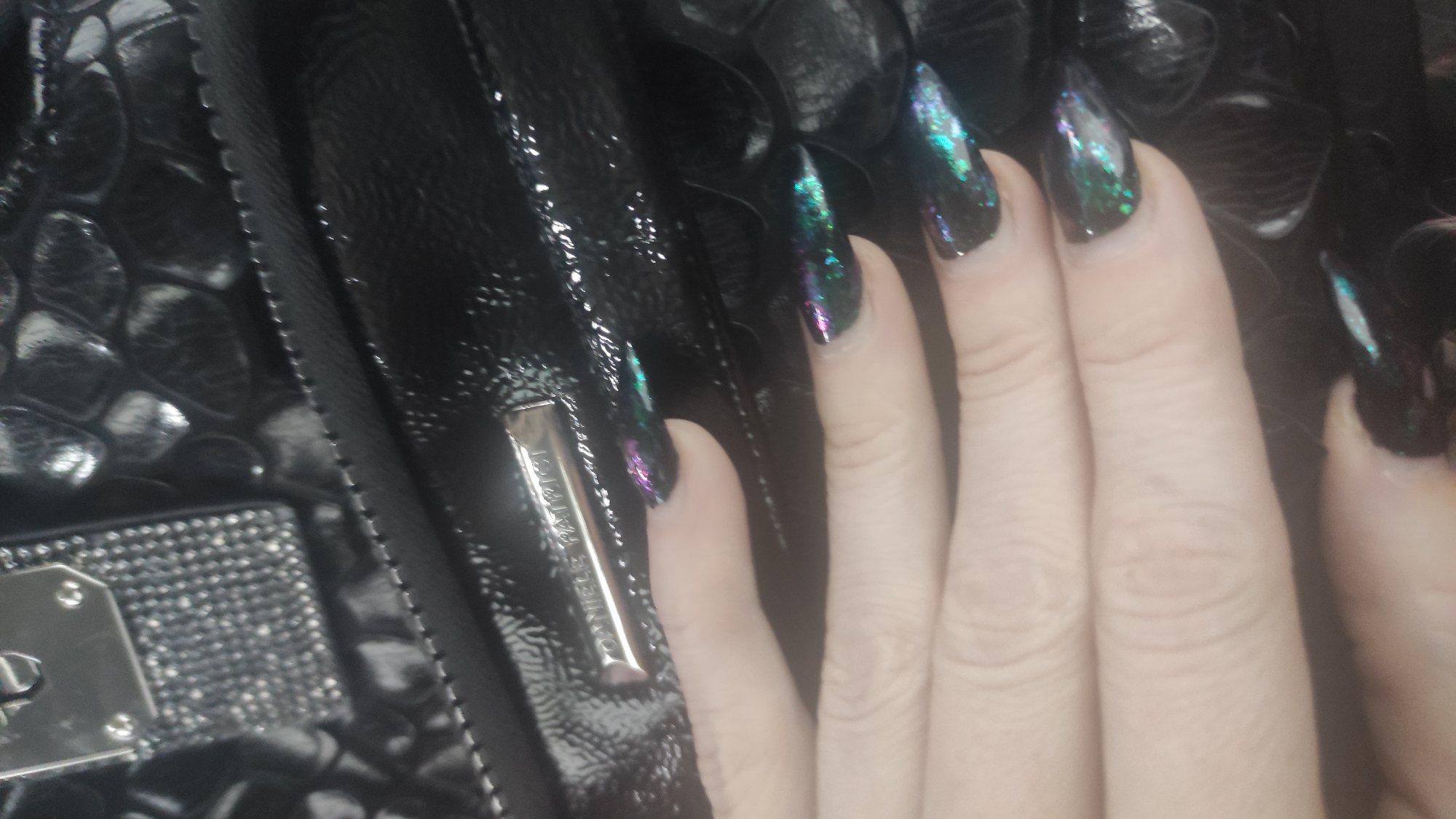 This close-up image, set in Chaikovsky, Russia, prominently features a human hand with long, pointed nails. The nails are painted with a dark base and an iridescent or holographic glitter overlay, reflecting vibrant green, purple, and blue hues. The hand rests on a glossy, black accessory, which appears to be a patent leather handbag or wallet with a textured pattern resembling crocodile or alligator skin.
A silver-colored metal bar, likely a zipper pull or decorative element, is visible on the bag. Partially engraved on this metal bar is the text "DANIELS PATRIC". Another section of the bag features a detailed pattern of small, sparkling elements, possibly studs or rhinestones, adding to its embellishment. The lighting emphasizes the reflective surfaces of both the nails and the bag, highlighting their textures and finishes. The scene appears to be an indoor setting, likely focusing on showcasing the stylish accessories. No specific activity or event beyond posing for the photograph is depicted, and no particular emotions are conveyed.