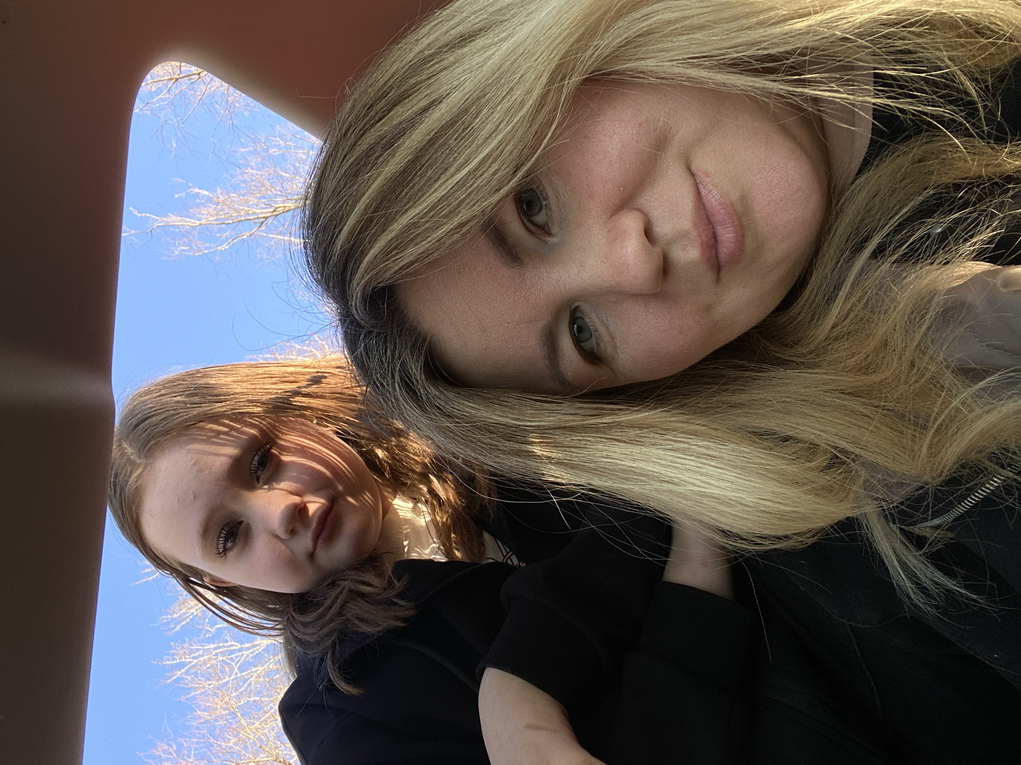 This image, taken from a low, tilted angle, features two females, likely inside a vehicle, with a clear blue sky and bare trees visible in the background.
The primary subject, in the foreground, is an older female with long, flowing blonde and brown streaked hair that frames her face. She has light-colored eyes and a neutral, calm expression, looking directly at the viewer with a hint of a soft smile. Her skin appears smooth, and she wears what looks like a dark, possibly black, jacket or top, with visible fabric texture.
Behind and to the left of the foreground subject is a younger female. Her long, dark brown hair is visible, and her face is partially covered by distinct shadows, creating a striped pattern that suggests light filtering through bare branches or a car's interior design element. She also has a neutral, composed expression as she gazes towards the camera. She is similarly dressed in a dark garment.
The scene outside the vehicle, viewed through a window or sunroof, reveals a bright, cloudless blue sky. Against this vibrant backdrop, the intricate, delicate silhouettes of bare tree branches are prominent, indicating a season like late autumn, winter, or early spring. The bright sunlight suggests it is daytime with clear weather. Parts of the vehicle's interior trim, likely the ceiling or a pillar, frame the outdoor view on the left side of the image.
The activity appears to be a casual moment, perhaps a selfie or a spontaneous portrait, captured while the subjects are traveling or resting in a car in Chaikovsky, Russia. No visible text is present in the image.