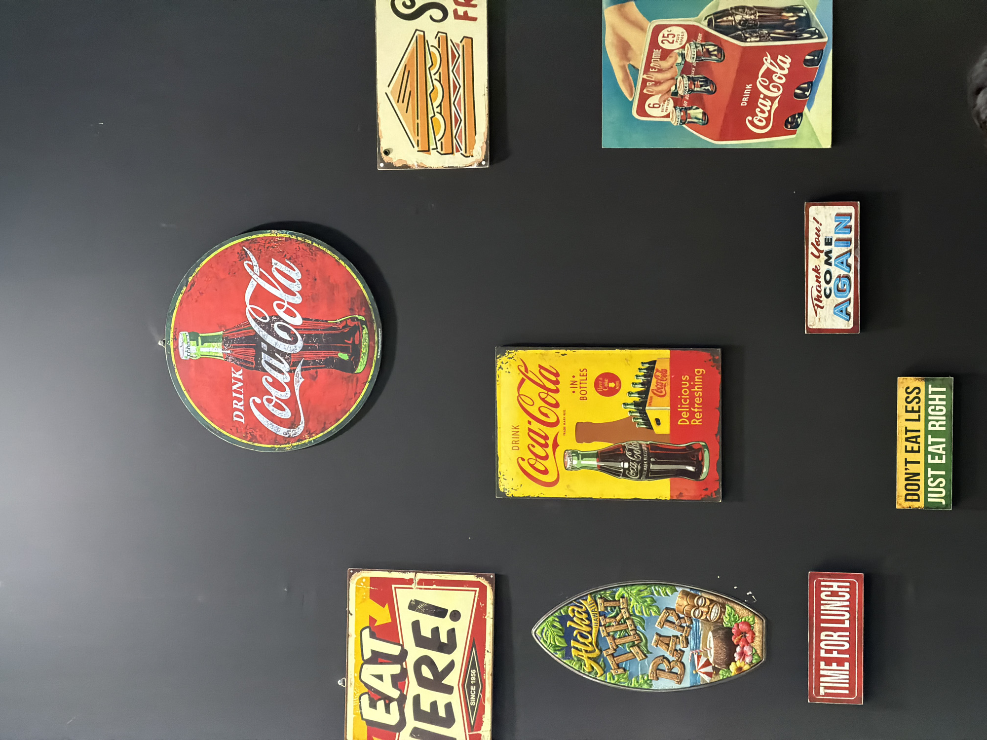 A collection of vintage-style signs are displayed on a black wall, possibly in a restaurant or bar in Kuta, Indonesia. The signs feature retro advertisements and slogans related to food and drinks, with a prominent Coca-Cola theme.
The signs include:
* A circular red Coca-Cola sign with a bottle and the text "DRINK Coca-Cola".
* A rectangular yellow sign with red lettering that reads "EAT HERE! SINCE 1956".
* A vertical yellow sign with red accents, also advertising Coca-Cola, featuring a bottle and text stating "DRINK Coca-Cola... IN BOTTLES... Delicious Refreshing".
* A surfboard-shaped sign with a tropical scene and the text "Aloha Tiki BAR".
* A rectangular red sign with white lettering that reads "TIME FOR LUNCH".
* A rectangular yellow sign with green lettering that reads "DON'T EAT LESS JUST EAT RIGHT".
* A rectangular sign with white lettering on a red background that reads "Thank You! COME AGAIN".
* A rectangular sign depicting a sandwich, with the text "S... FR...".
* A rectangular sign featuring a hand holding a six-pack of Coca-Cola bottles, with pricing information.
The black wall provides a neutral backdrop, making the colorful signs stand out. The overall impression is one of a themed establishment that embraces nostalgia and classic Americana.