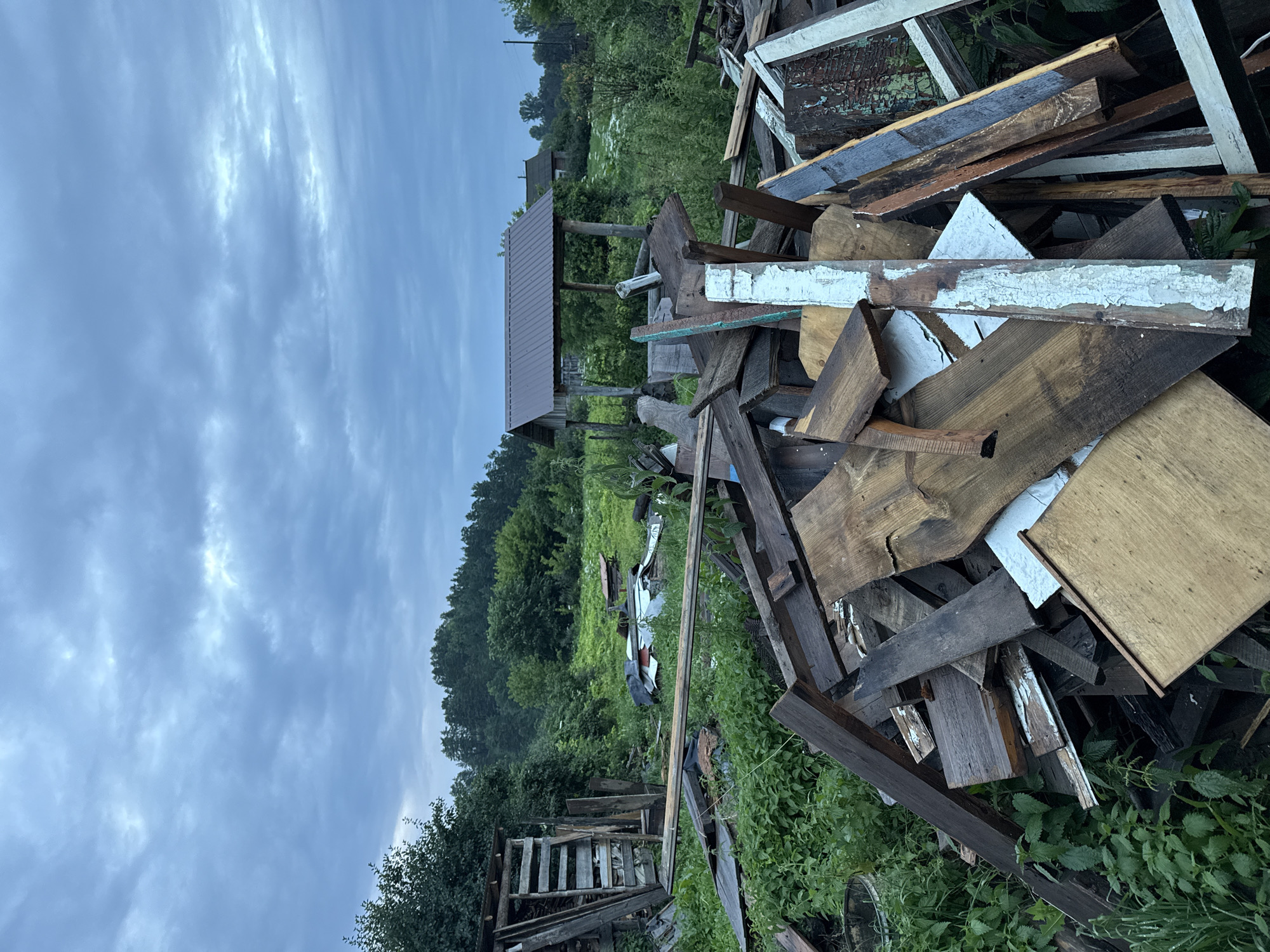 In a rural outdoor setting, likely Gon'ba, Russia, a large pile of discarded lumber and building materials dominates the foreground. The materials are assorted in shape, size, and condition, with some pieces appearing weathered and others newer. The lumber is densely packed and jumbled, creating an uneven terrain within the frame.
Behind the pile of debris, the environment transitions to a natural landscape. Lush green vegetation, including tall grass and bushes, surrounds the lumber. In the mid-ground, a simple, rustic wooden structure with a metal roof stands to the right, possibly a shelter or wellhead. Further in the background, a dense line of trees forms a treeline against a cloudy, overcast sky. The overall impression is one of neglect or a site undergoing demolition or construction cleanup in a rural area. No people or animals are visible in the image. The lighting suggests it is late afternoon or early evening, or the day is heavily overcast.