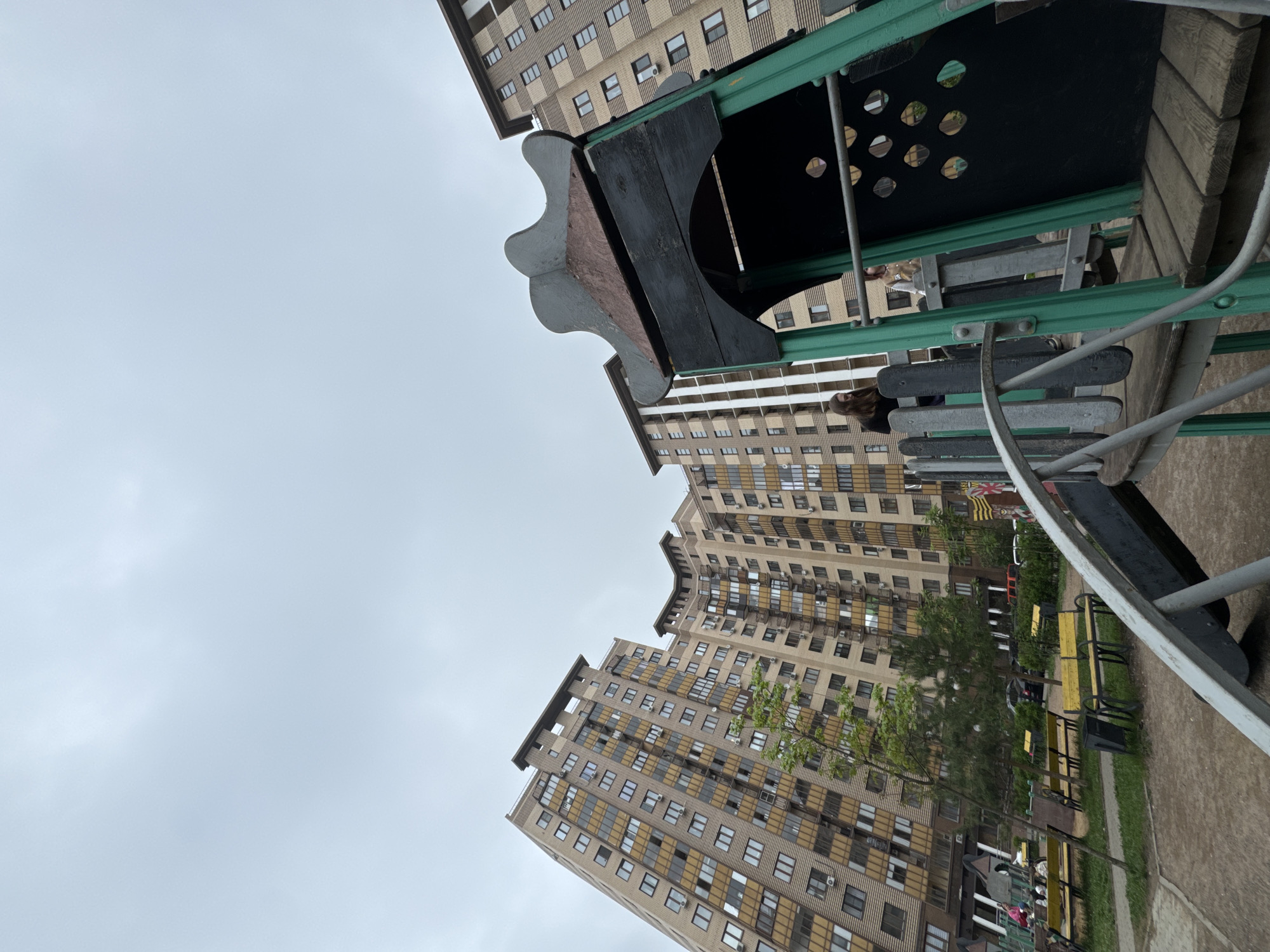 The image shows a view of an outdoor area, likely a playground or park in Odintsovo, Russia. In the foreground is playground equipment with a climbing structure made of metal and wood/plastic components. The playground is positioned in a manner which makes the frame seem upside down. In the background, tall apartment buildings with many windows and balconies are visible. These buildings form a backdrop to the park. The sky is overcast. The playground is located among the buildings, with benches and trees.