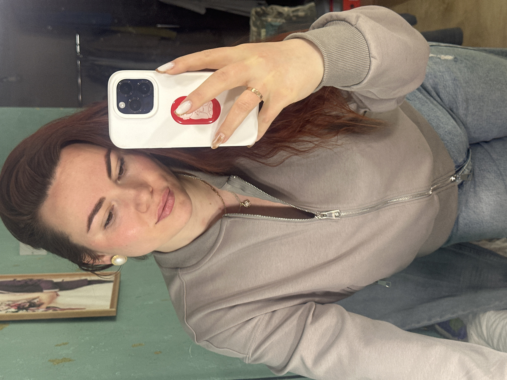 A woman is taking a selfie in what appears to be a room or apartment, possibly in Черёмушки, Russia, although this cannot be definitively confirmed without additional information or context.
The woman has reddish-brown hair and is wearing a light gray zippered jacket, a delicate necklace, and a pearl earring. She is holding a white smartphone with a distinctive red and white design on the case, and a gold ring is visible on her finger. She has light skin and is wearing makeup.
She is wearing blue jeans with what appear to be distress marks. Behind her and the woman in the frame is a painting on a light green surface. Parts of furniture/bedding are visible in the background. A reflection is seen in the top left corner.