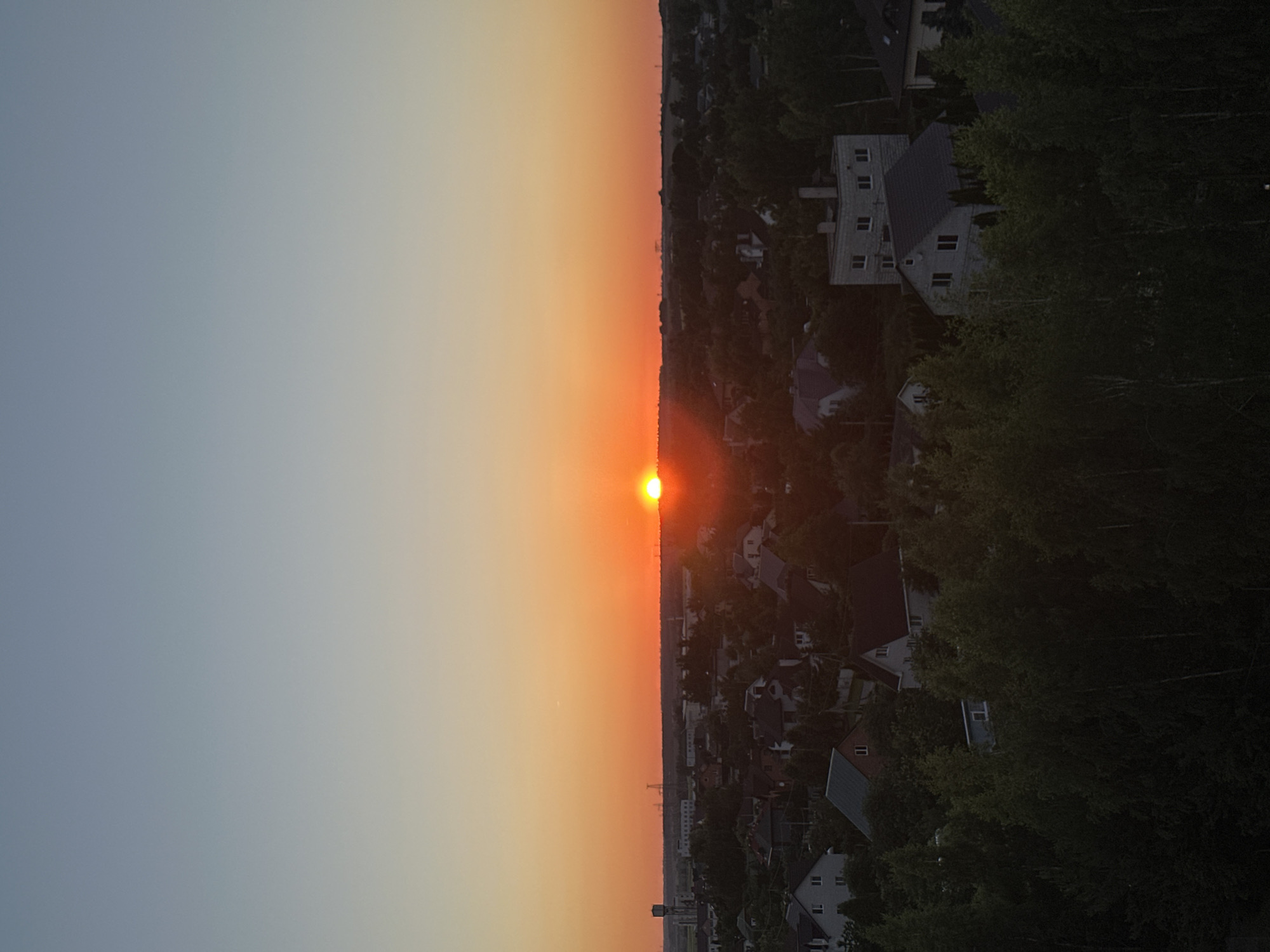The image captures a sunset over a residential area. The sun, a bright orange orb, is positioned just above the horizon line, casting a warm glow across the sky. The sky transitions from a pale blue at the top to a vibrant orange near the horizon. Below the sky, a landscape of houses is visible, nestled among lush green trees. The architecture of the houses appears varied, with some featuring pitched roofs and others more modern designs. The foreground is dominated by dense foliage, suggesting a natural setting within or on the outskirts of a populated area. The overall scene evokes a peaceful, twilight ambiance. This view is consistent with locations in the Шчомысліцкі сельскі Савет, Belarus, a region characterized by rural and suburban settlements with ample green spaces.