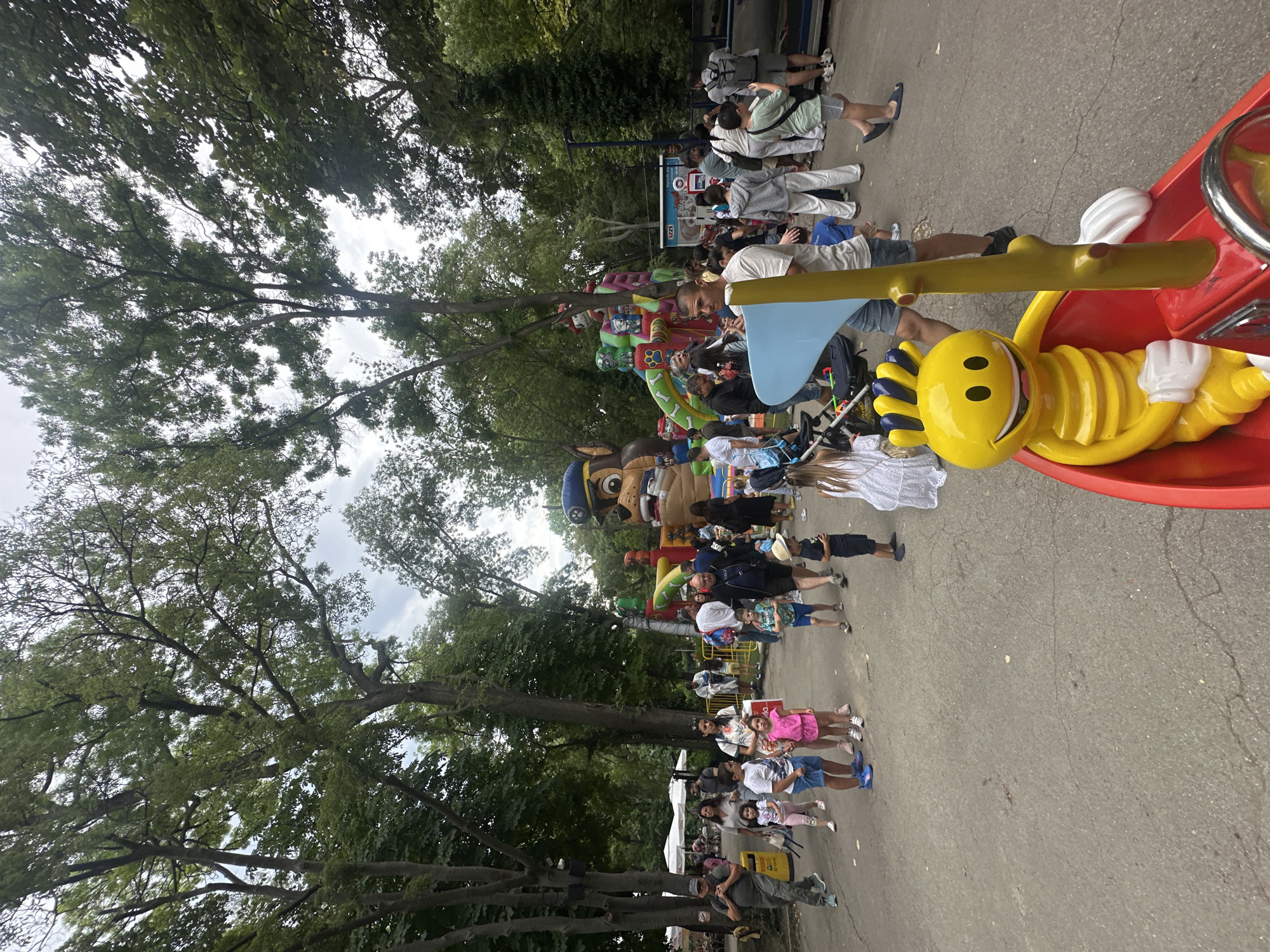 In a park setting with large trees overhead, a crowd of people, including adults and children, are gathered. A large inflatable structure, featuring characters from Paw Patrol, is visible in the background. Several people are walking along a paved pathway. In the foreground, a yellow cartoon character figure is seated in a red boat-like structure. The scene suggests a public event or fair.