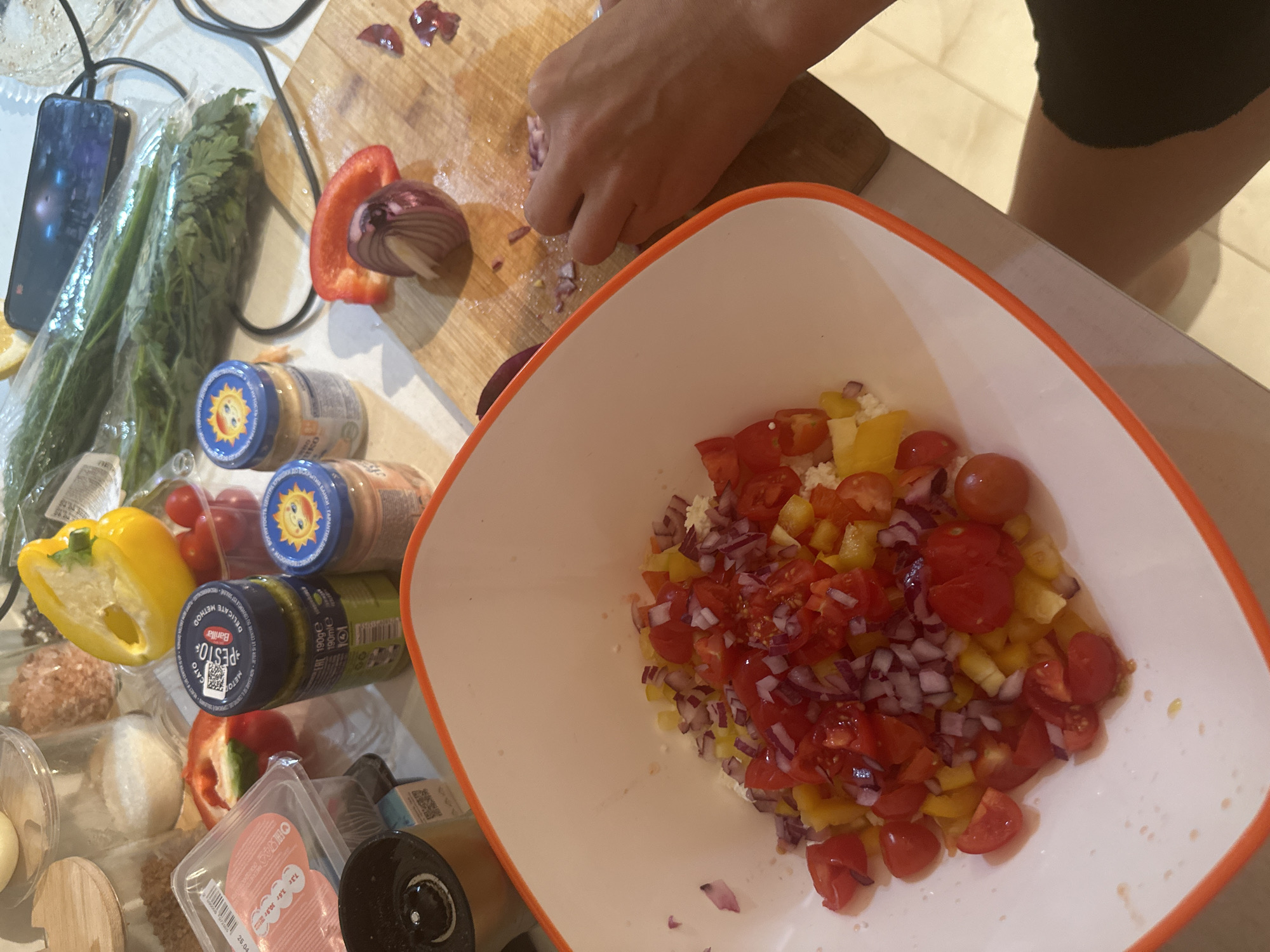 The image captures a top-down view of a kitchen countertop where food preparation is underway. A person’s hands are visible at the center-top, using a knife to finely chop a red onion on a wooden cutting board. A red bell pepper half is also on the board.
To the right, a large white bowl with an orange rim contains a mix of diced ingredients: yellow bell pepper, cherry tomatoes, and red onion, resting on a base of what appears to be crumbled white cheese or a similar ingredient.
The countertop is cluttered with various food items and kitchen essentials. On the left, a smartphone is plugged in and rests next to a plastic bag containing fresh green herbs, possibly parsley or dill. Below these are several jars, including three baby food jars with a cheerful sun logo on their lids. One jar is clearly identifiable as "Barilla PESTO" with "METODO DELICATO" written on its label, along with nutritional information "7,3г 3,6г 30,9г" and a date "25.04". Further left, a yellow bell pepper half and a container of what looks like ground meat or meatballs are visible, alongside a clear plastic container possibly holding eggs or mozzarella balls. Another red bell pepper half and a black spice grinder are also present on the cluttered surface.
The activity clearly depicts the process of assembling a fresh meal, likely a salad or a salsa. The setting is an indoor kitchen, suggested by the countertop, cutting board, and various ingredients.