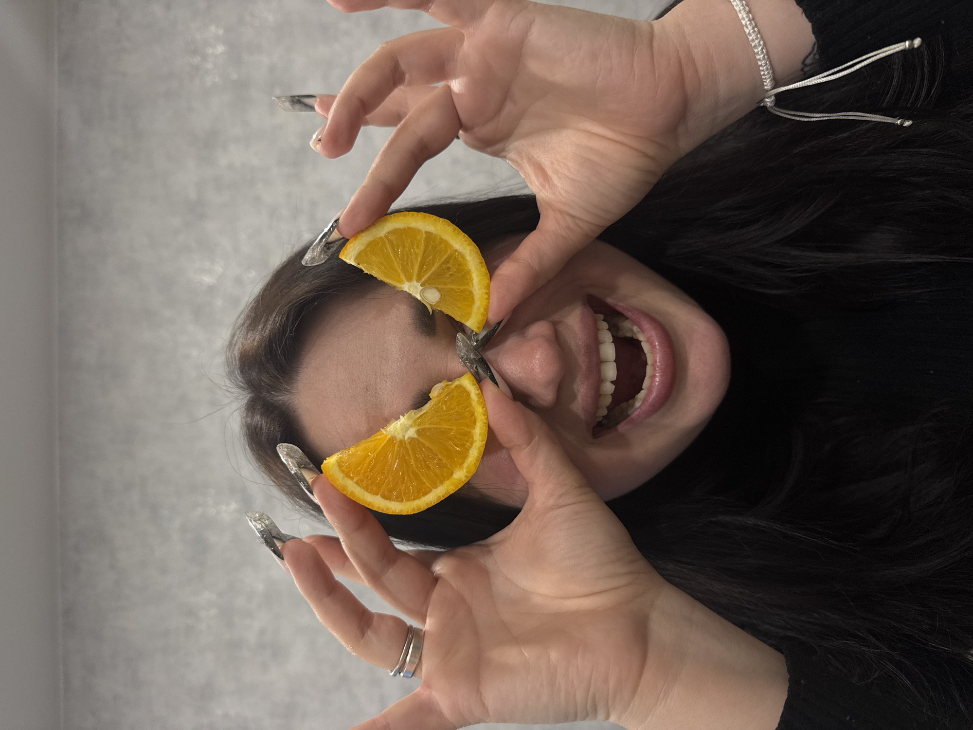 An eye-level shot captures a person, likely female, with long, dark hair, playfully holding two bright orange slices over their eyes. The image is oriented vertically, with the person's head tilted sideways, and they are looking upwards with their mouth wide open, revealing teeth and tongue, suggesting a moment of laughter or a playful expression.
The person's hands are prominent, adorned with long, pointed, and clear or silver-toned artificial nails. On their right wrist, a silver-colored braided or macrame-style bracelet is visible. Two simple silver rings are worn on the ring finger of the left hand. One of the orange slices, positioned over the left eye, clearly shows a small seed. The background is a plain, light grey, slightly textured wall, indicating an indoor setting. The overall mood of the image is one of lightheartedness and fun. There is no visible text in the image.