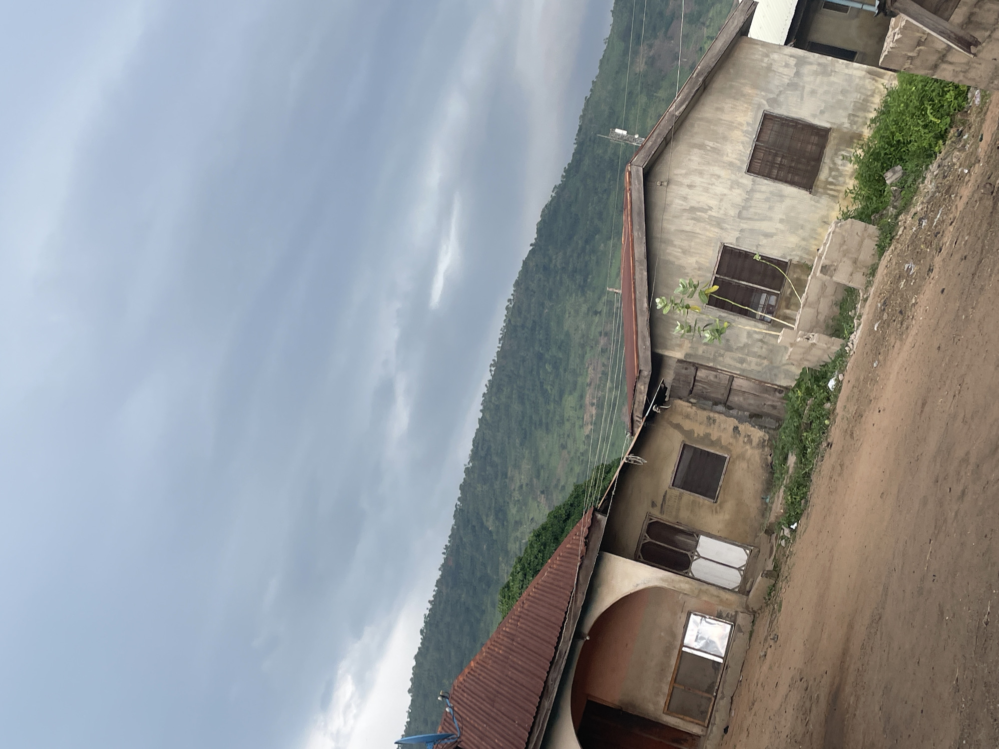 The image depicts a slightly tilted, overcast scene of two modest, weathered buildings in Lokoja, Nigeria. The structures appear to be residential, with corrugated metal roofs, some in disrepair. The building on the left has a reddish-brown, rusted corrugated metal roof and an arched doorway with a white door. It also has a window with a reflective surface. The building on the right is a lighter gray, with a similarly sloped corrugated metal roof. It features two windows with dark, slatted coverings and is adjacent to a more primitive structure.
The ground in front of the buildings is a dusty, brown dirt road. Overgrown green vegetation is visible around the base of the buildings and between them. In the background, a lush, green, tree-covered hillside or mountain rises steeply against a cloudy, gray sky. The overall impression is of a rural or semi-rural setting, with a natural landscape dominating the background. No people are visible in the image.