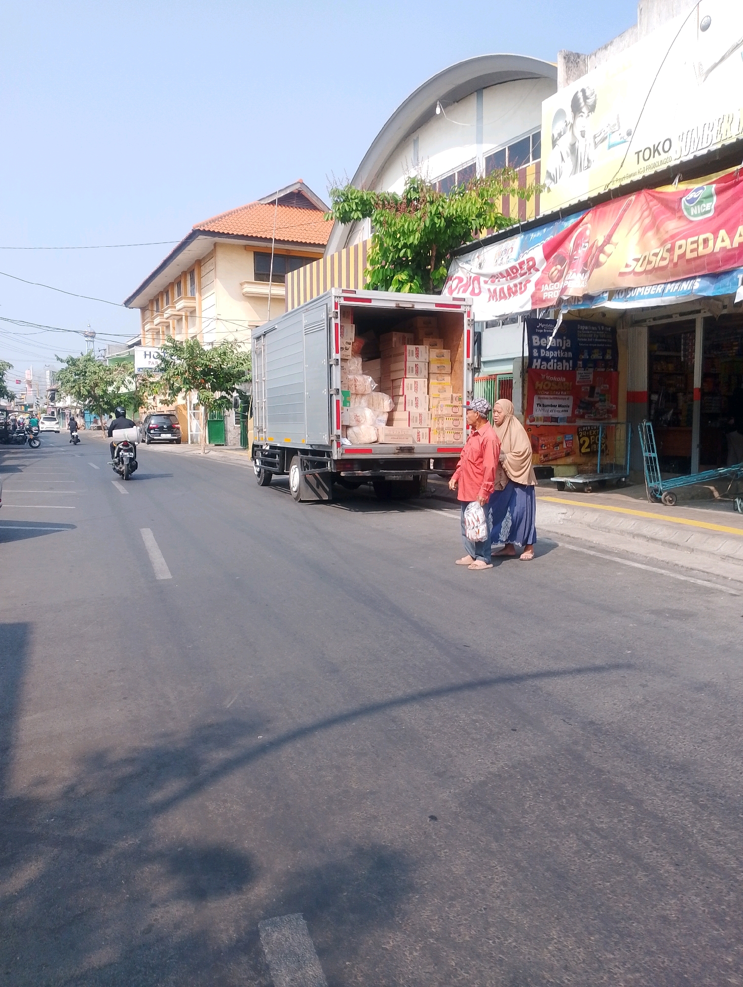 In Probolinggo, Indonesia, a street scene unfolds under a clear, pale blue sky. The sun casts long shadows across the asphalt, indicating it is likely mid-morning or late afternoon. A large, silver box truck, partially loaded with cardboard boxes, is parked on the right side of the road, its rear doors open. Two women, both wearing headscarves, stand on the sidewalk near the truck. The woman on the left is wearing an orange long-sleeved shirt and blue pants, holding a plastic bag. The woman on the right is in a light brown headscarf and blue skirt. They appear to be either interacting with the truck or observing its contents.
Several motorcycles are visible on the road. One motorcycle is parked on the left, and another is being ridden away from the camera by a person wearing a black jacket and helmet, carrying a rolled-up mat. In the background, a black car is parked on the side of the road, and further down the street, other vehicles and pedestrians are present.
The buildings lining the street include a multi-story building with a terracotta-tiled roof on the left, exhibiting a yellowish facade with multiple windows. On the right, a commercial building features a large awning with various advertisements, including one for "TOKO SUMBER MANIS". A narrower building with a red awning and a display of food items is also visible. The sidewalk is paved, and a blue hand truck is positioned near the commercial building. Trees with green foliage are interspersed along the roadside. The overall impression is a typical urban street scene in Indonesia, likely bustling with commercial activity.