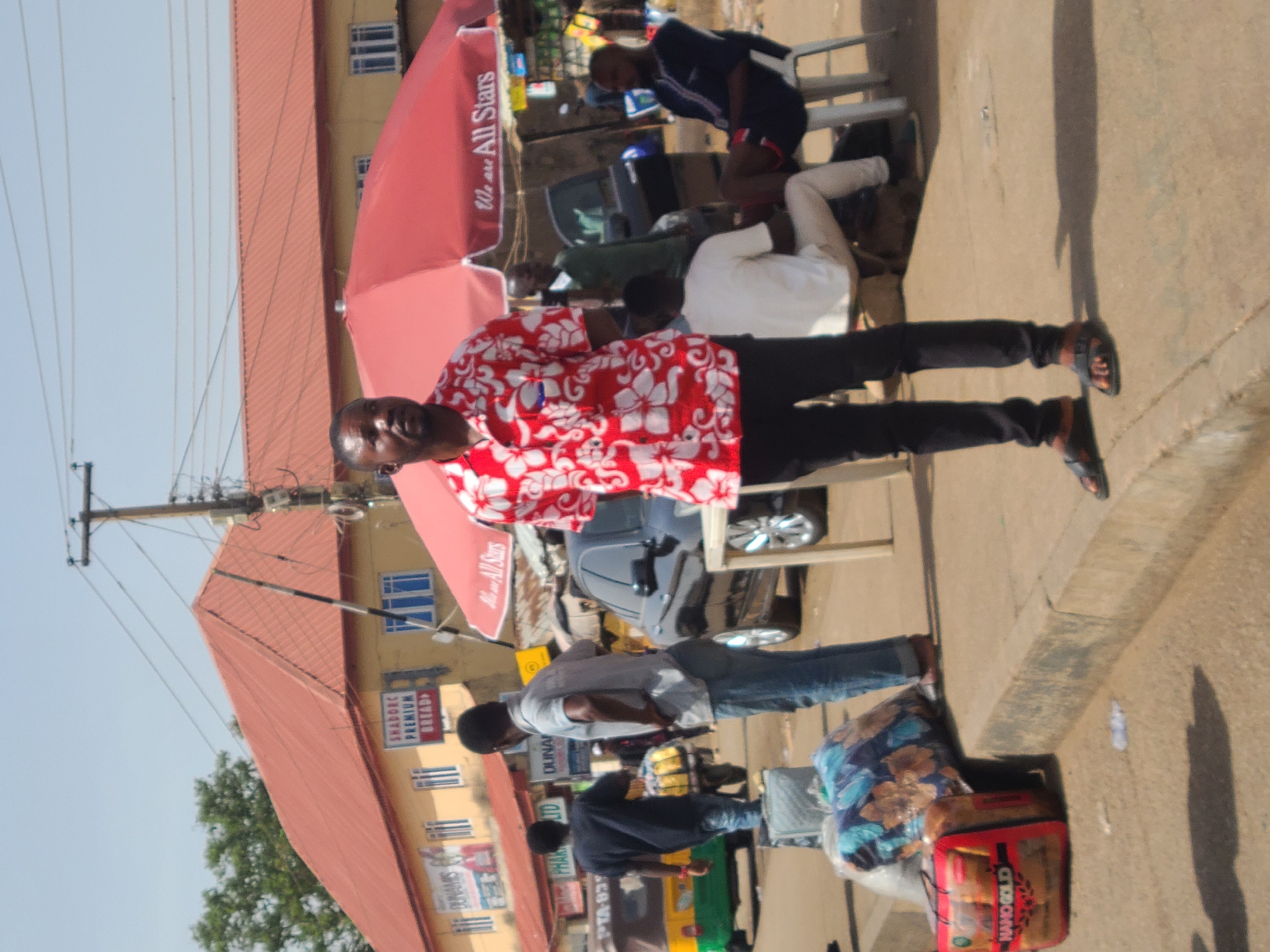 The image captures an urban scene in Jos, Nigeria, displaying a variety of activities. A man in a red and white floral shirt and black pants stands on the sidewalk, seemingly posing for the photo. In the background, a building with a red roof and cream-colored facade stands, adorned with signs, including one for "SHADREK PREMIUM BREAD". Street vendors and pedestrians populate the scene, with a tricycle taxi (Keke Napep) visible amongst other vehicles. One individual is seated under a red umbrella. Goods and luggage are piled on the pavement near a vendor. The overall environment suggests a bustling marketplace or commercial area in Jos. Overhead, power lines crisscross the sky.