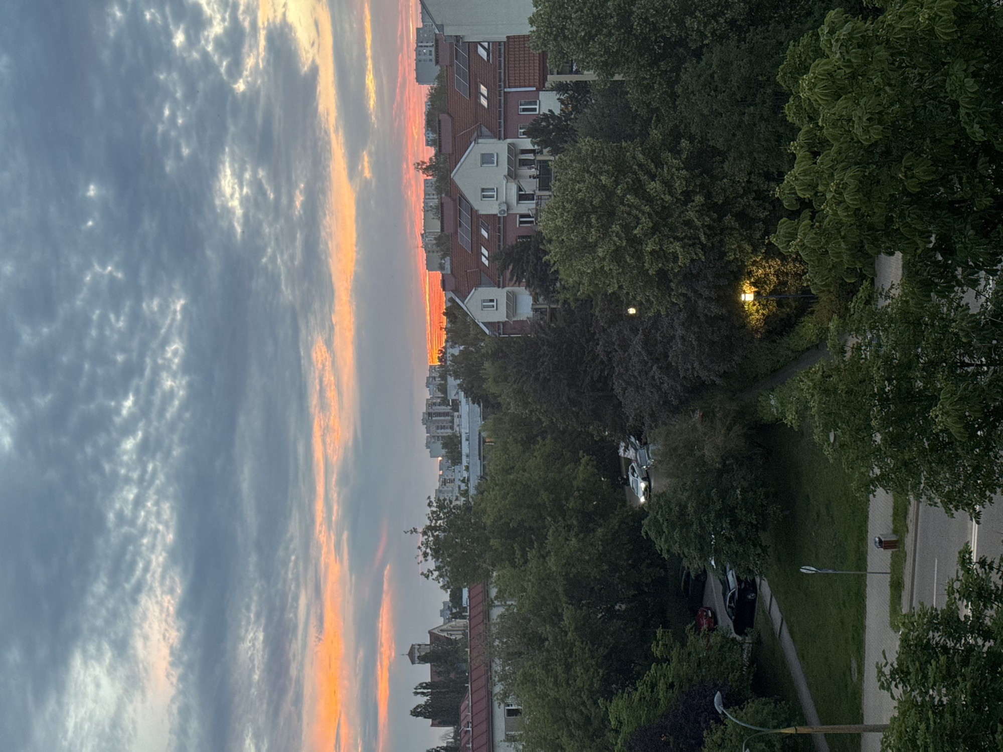 The image depicts a street scene in Warsaw, Poland, during what appears to be sunset or dusk. The sky is filled with dramatic clouds, painted with hues of orange and pink from the setting sun, contrasted with darker gray clouds. Below the sky, a row of buildings with red-tiled roofs and white facades stands, some featuring solar panels. Lush green trees line the street, with some overhanging the buildings and the pavement.
A paved pathway or sidewalk is visible, bordered by grass. Several cars are parked along the side of the street and on the sidewalk area. One white car with its headlights on is turning or driving on the road. A streetlamp with a visible light source illuminates a patch of the greenery. Another streetlamp is visible in the foreground, without its light on. The overall atmosphere suggests a residential urban environment at the end of the day. The orientation of the image is landscape but the content is rotated 90 degrees clockwise.