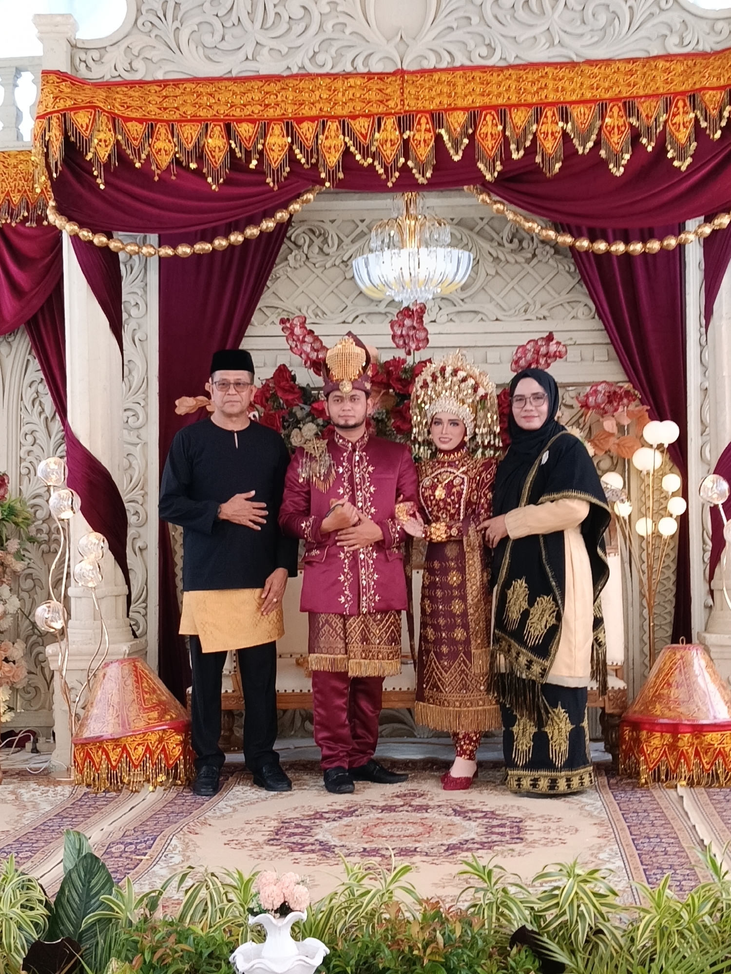 The image shows four individuals posing in front of a decorative backdrop, possibly at a wedding venue in an unknown city. The backdrop features burgundy drapes, a beaded garland, and an ornate white carved panel. The floor is covered in a patterned carpet, and there are plants along the bottom edge of the image.
From left to right:
1. A man wearing a black long-sleeved shirt, black pants, and a yellow sarong. He has short dark hair and is wearing glasses. His left hand is placed on his stomach.
2. A man wearing a maroon and gold patterned suit and a traditional headdress. He is holding his hands clasped in front of him.
3. A woman wearing a maroon and gold patterned dress with a tall, ornate headdress.
4. A woman wearing a black hijab, glasses, and a light-colored garment covered with a black patterned shawl. Her left arm is extended, and her hand is resting on the arm of the bride.
The scene is illuminated by the overhead chandelier and the decorative lamp fixtures on both sides.