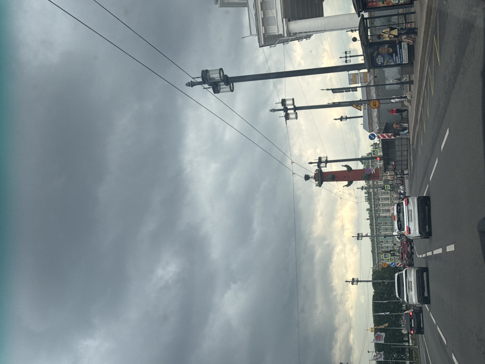 This is a street-level, eye-level, forward-facing shot captured from a vehicle, looking down a multi-lane road in Saint Petersburg, Russia. The sky is overcast with heavy, dark clouds. The road has clear lane markings and is populated with several vehicles, including white SUVs, a black sedan, and other cars further ahead. On the right side of the road, there is a bus stop with people waiting and advertisements displayed. Streetlights line the road, and to the left, there are flags and trees. In the background, an ornate, long, light-green building with white columns and windows stands prominently. In the middle ground, a tall, red Rostral Column, a historical landmark in Saint Petersburg, is visible. The overall scene depicts a busy urban environment under a cloudy sky.