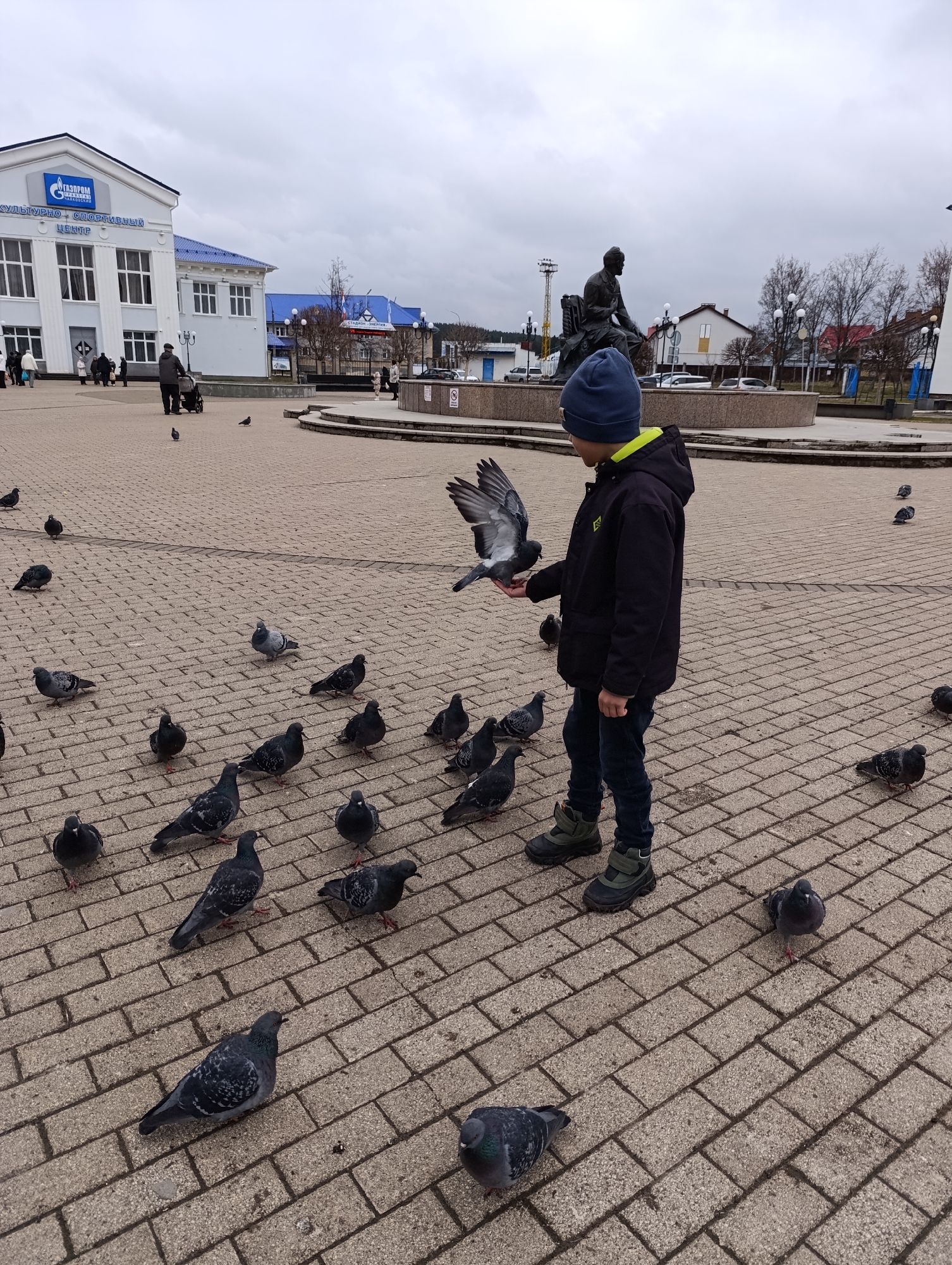The image depicts an overcast daytime scene in a public square in Chaikovsky, Russia. The central focus is a young boy, wearing a dark jacket, blue hat, jeans, and sturdy boots, standing on a paved surface. He is actively interacting with pigeons, with one pigeon perched on his hand, appearing to be fed. A large number of other pigeons are scattered on the ground around him, some very close, suggesting a common feeding spot.
In the background, to the left, stands a prominent light-colored building with a blue roof and classical columns. Visible text on this building identifies it as "ГАЗПРОМ КУЛЬТУРНО - СПОРТИВНЫЙ ЦЕНТР" (Gazprom Cultural - Sports Center), featuring the Gazprom logo. Several adults are walking near this building, one pushing a pram. To the right of the square, a dark statue of a seated figure overlooks a circular fountain structure. Further beyond, bare trees suggest a cooler season, and additional buildings are visible, including one with a "СТАДИОН ЭНЕРГИЯ" (Stadium Energiya) sign. The square is paved with light grey interlocking bricks. The weather is uniformly cloudy, indicating a cool and likely muted light conditions.