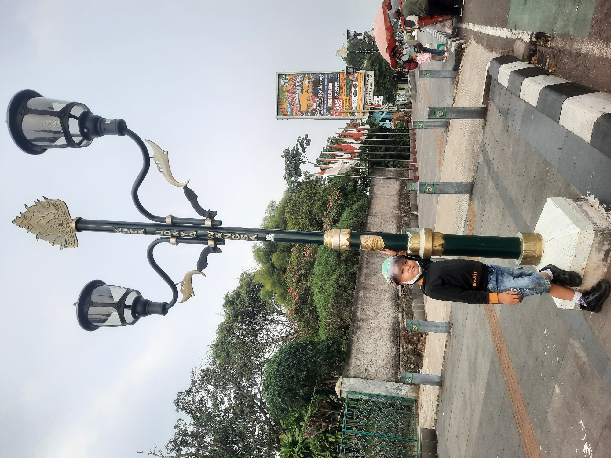 In Sukaraja, Indonesia, a young boy wearing a helmet and a black hoodie with orange trim, along with denim shorts, stands leaning against a decorative lamppost. The lamppost features multiple lights and ornamental gold accents. The boy is positioned on a paved walkway with a black and white striped curb to his right. In the background, trees, a stone wall, a green fence, several Indonesian flags, and a billboard advertising a festival are visible. The sky is overcast.