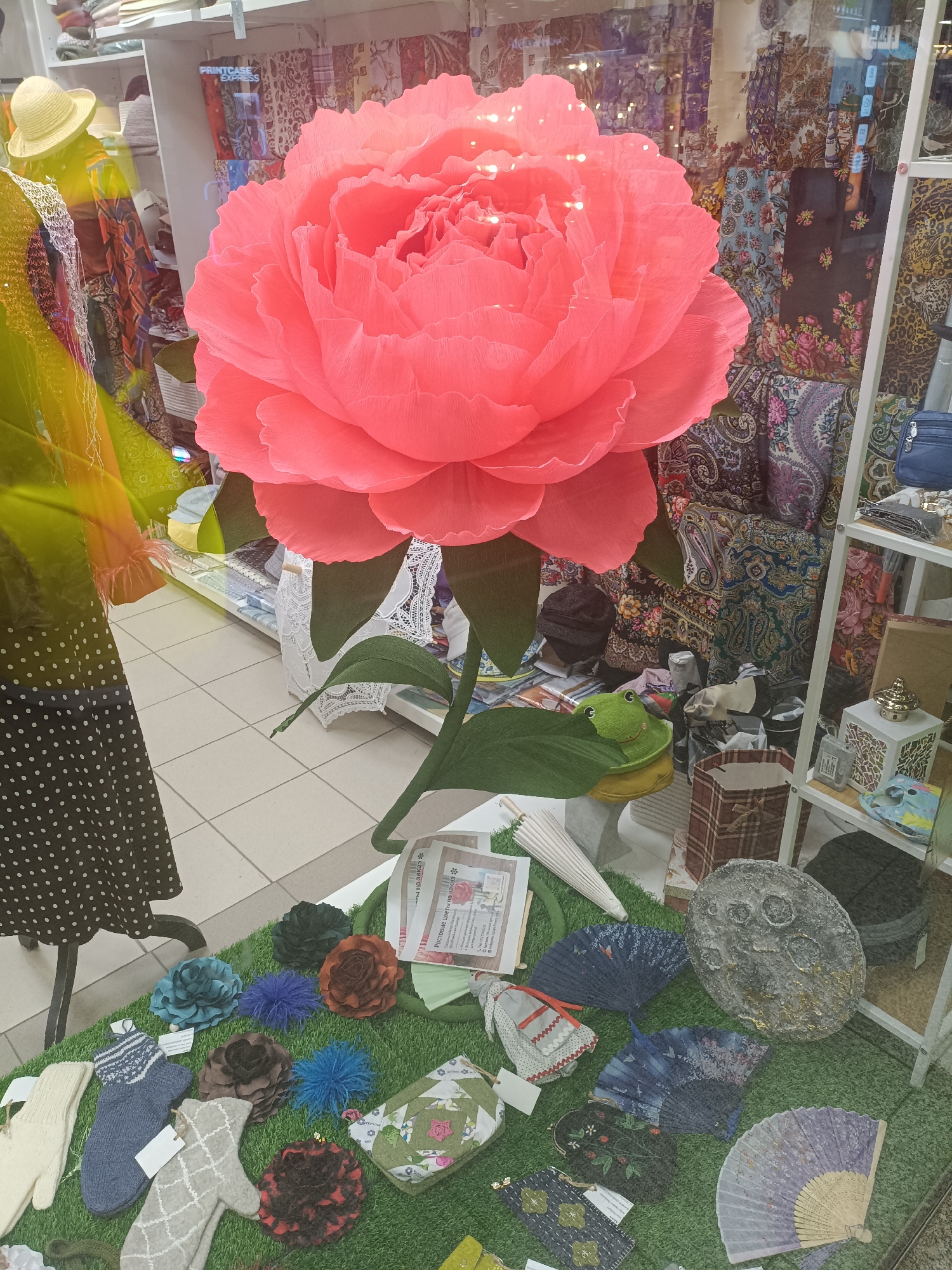 This is a photograph of a shop window display in Narva, Estonia. The display features a large, artificial pink peony flower as the central element. To the left of the flower, a mannequin is dressed in a black polka-dot dress and a yellow hat. Behind the flower and mannequin, the shop's interior is visible, showcasing shelves filled with textiles, scarves, and other merchandise. In the foreground, arranged on a patch of artificial grass, are various decorative items including knitted mittens, fabric flowers, small fans, a patterned pouch, and a textured, grey, moon-like object. A small, green stuffed frog sits on a large green leaf, adding a whimsical touch to the display. The floor of the shop appears to be tiled. The overall impression is one of a retail environment, likely a gift shop or textile store, presenting its wares in an attractive window display.