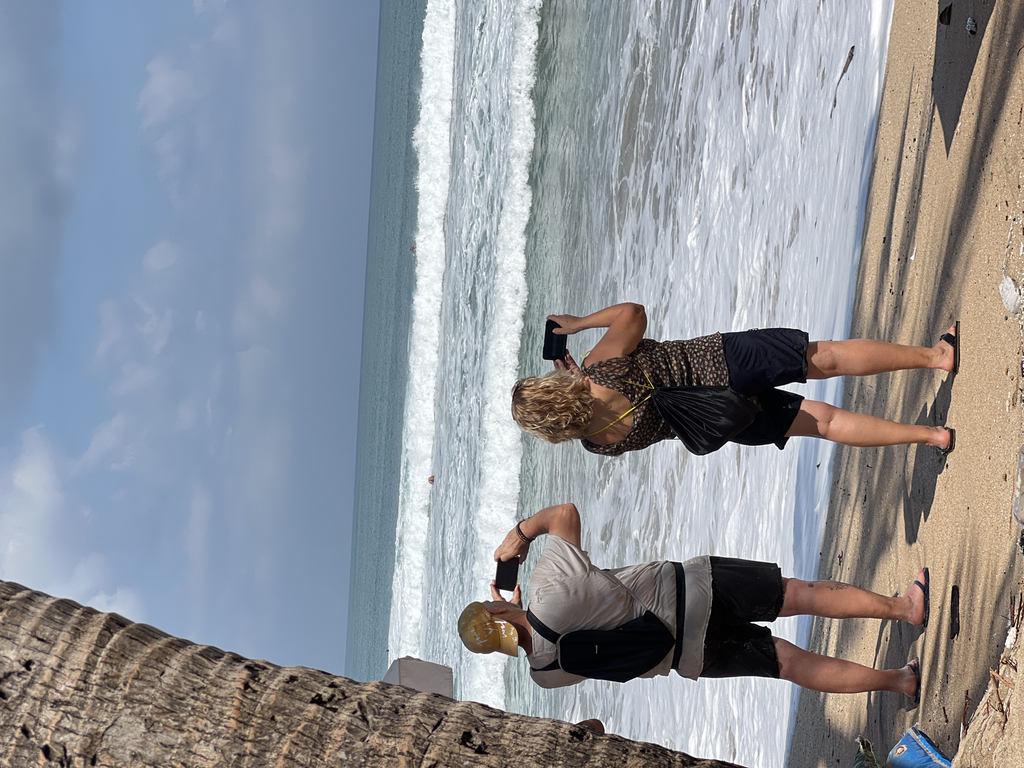 In Kuta, Indonesia, a sandy beach is populated by two individuals. A man, wearing a tan cap, a light-colored t-shirt, and dark shorts, stands with his back to the viewer, holding a smartphone to capture a photograph of the ocean. He also wears a black backpack and sandals. To his right, a woman, with blonde curly hair, is also facing the ocean and holding a smartphone, taking a photo. She is dressed in a patterned top and dark shorts, and also carries a black backpack. Both individuals are positioned near the water's edge, where waves are breaking on the shore. The ocean is a vibrant turquoise, with white foam from the waves. The sky above is blue with scattered clouds. A large palm tree trunk is visible on the left side of the frame, partially obscuring the view. Debris, including a blue plastic bag and a bottle, is scattered on the sand in the foreground. In the distance, a person can be seen swimming in the ocean.