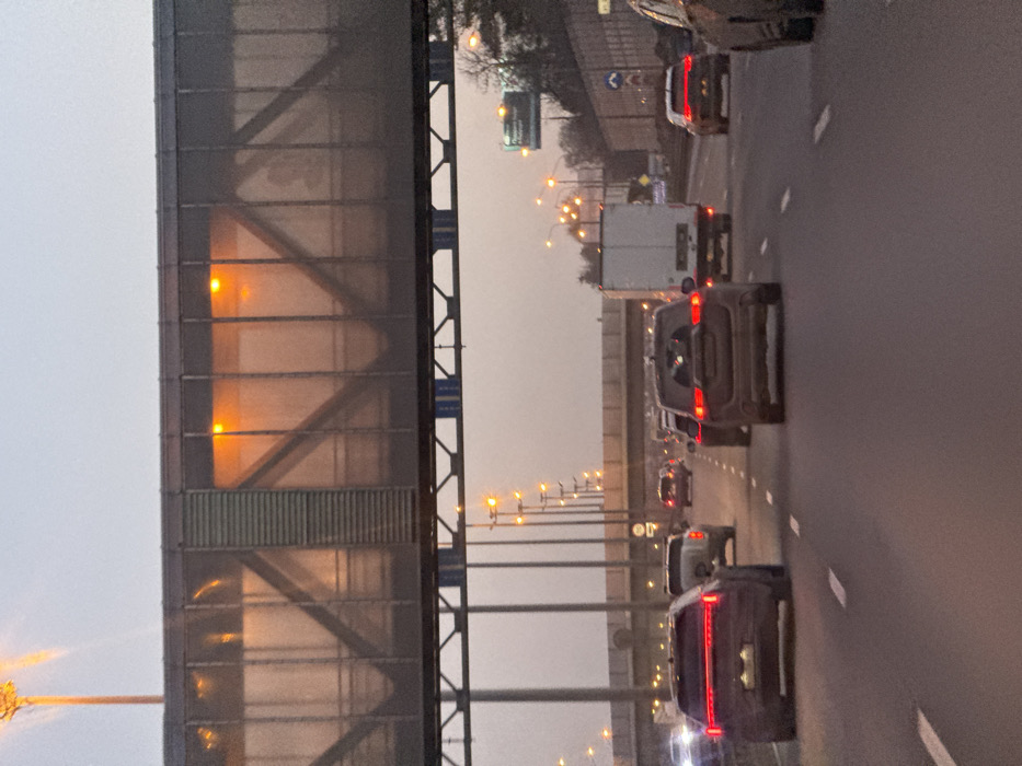 The scene depicts a multi-lane highway or major road in the city of Cheliuskinskii, Russia, during what appears to be dusk or dawn. The sky is a pale greyish-pink.
In the foreground, the view is from a vehicle traveling in the middle lane. Multiple vehicles are ahead, including a dark SUV in the leftmost lane with illuminated horizontal taillights, a dark SUV directly ahead in the middle lane also with illuminated taillights, and a white box truck in the right lane. Other vehicles are visible further down the road in all lanes, all with illuminated taillights. The road surface is dark asphalt with white dashed lane markings.
An overhead pedestrian bridge, constructed with a dark metal truss framework and transparent or translucent panels, spans the entire width of the highway. Inside the bridge, multiple orange-glowing lights are illuminated, casting reflections on the panels. Structural supports for the bridge are visible below.
Beyond the immediate overhead structure, a series of tall streetlights with warm orange lamps line both sides of the highway, extending into the distance. In the far background, another overpass or bridge crosses perpendicular to the main road. To the right of the highway, an elevated embankment is visible with sparse, bare trees and a partially obscured large green overhead sign.