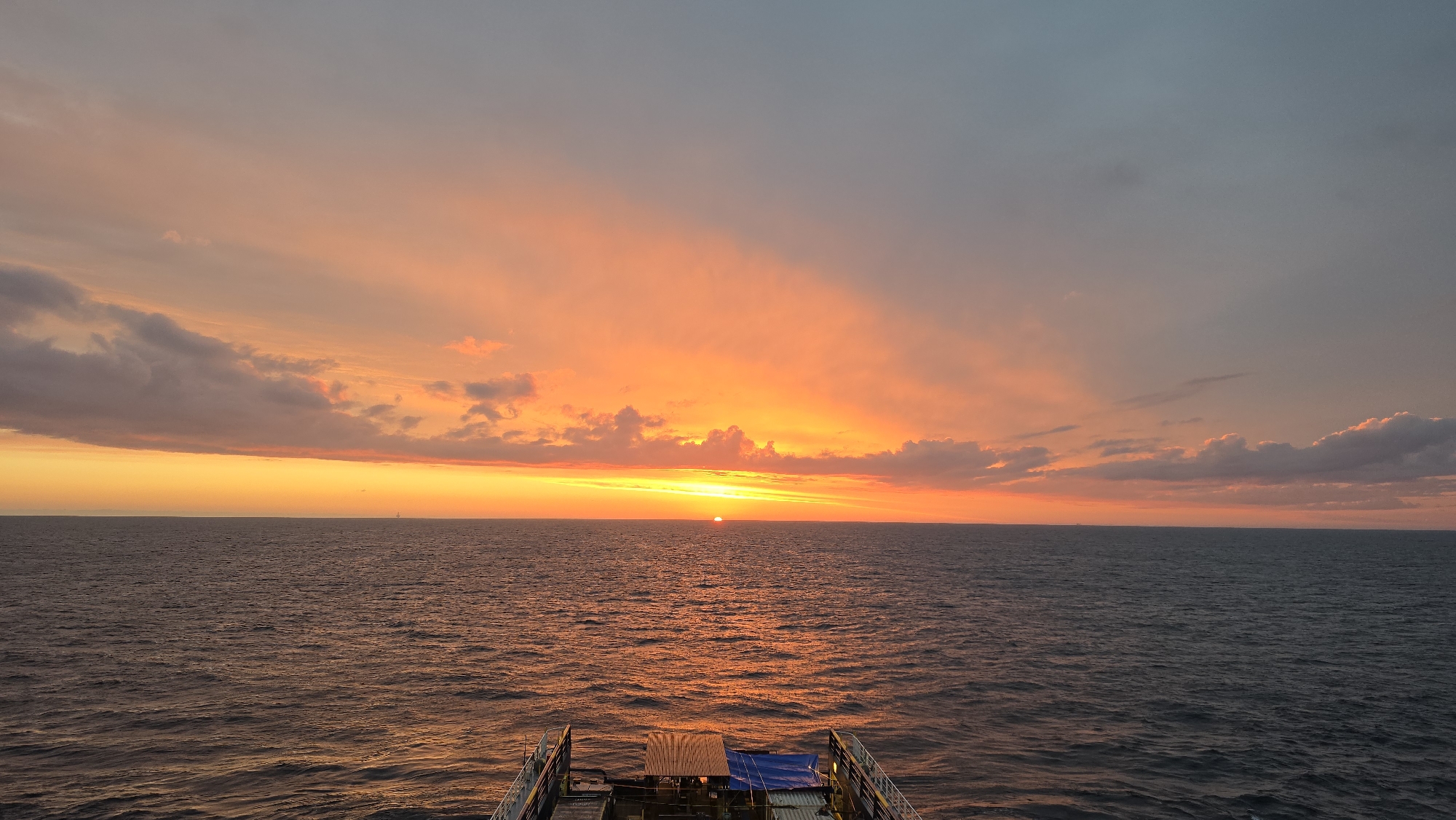 The image depicts a sunset over the ocean, viewed from the deck of a ship. The sky is filled with vibrant colors of orange, yellow, and pink, transitioning to a muted gray-blue higher up. The sun is low on the horizon, reflecting intensely off the water. The ocean surface is rippled, with a dark blue hue. The deck of the ship is visible in the foreground, with railings and some equipment. The location is the ocean near the city of Unknown, Unknown.