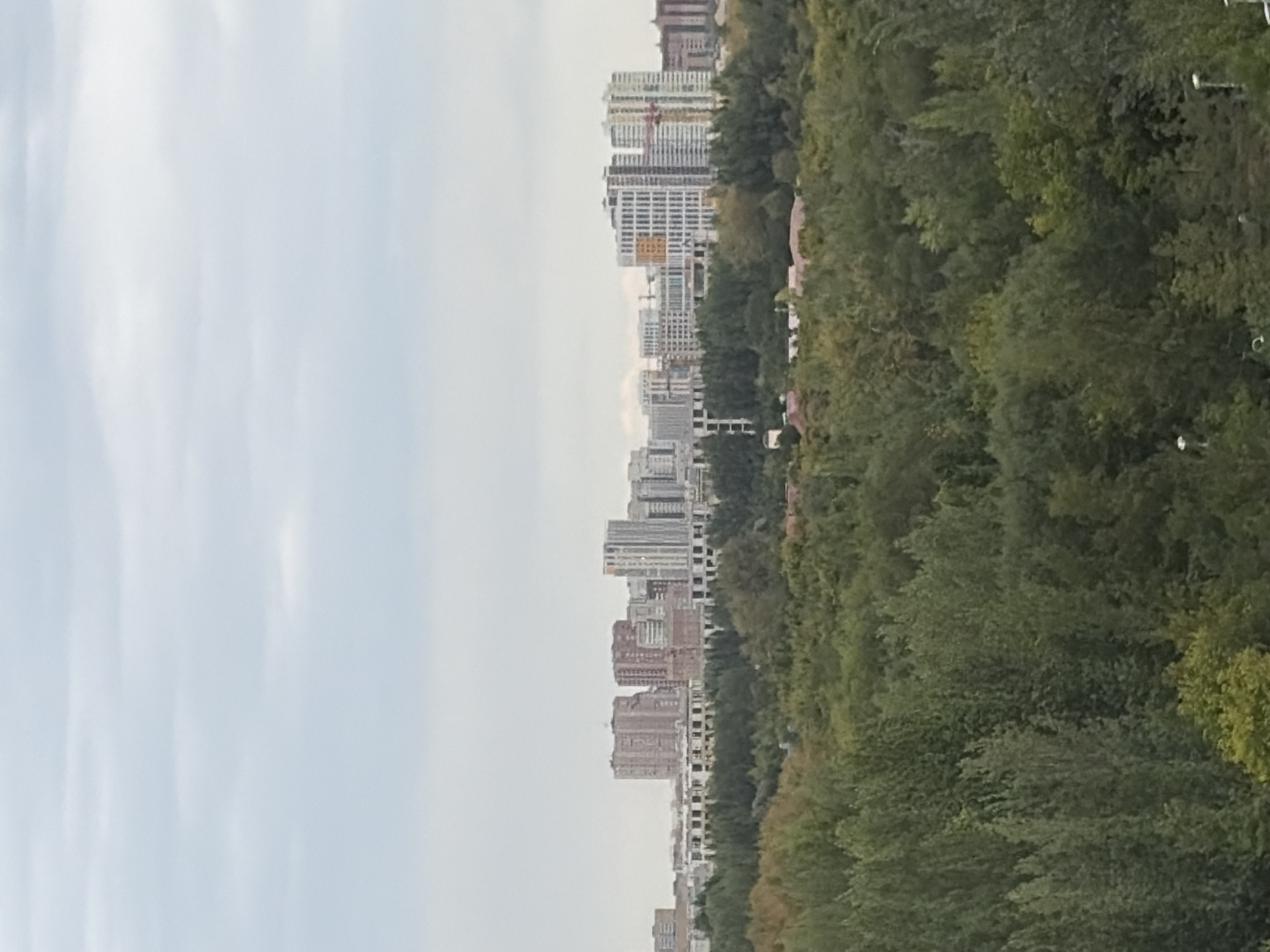 The image depicts a panoramic view of the cityscape of Astana, Kazakhstan, under an overcast sky. The foreground is dominated by a dense canopy of green trees, suggesting a park or forested area within the city. Beyond the trees, a sprawling urban landscape unfolds, characterized by a variety of modern buildings, including high-rise apartment complexes and other multi-story structures. Some buildings appear to be under construction, indicated by visible cranes. The architecture is contemporary, with a mix of glass facades and concrete structures. The overall impression is of a developing urban environment with a significant green buffer zone.