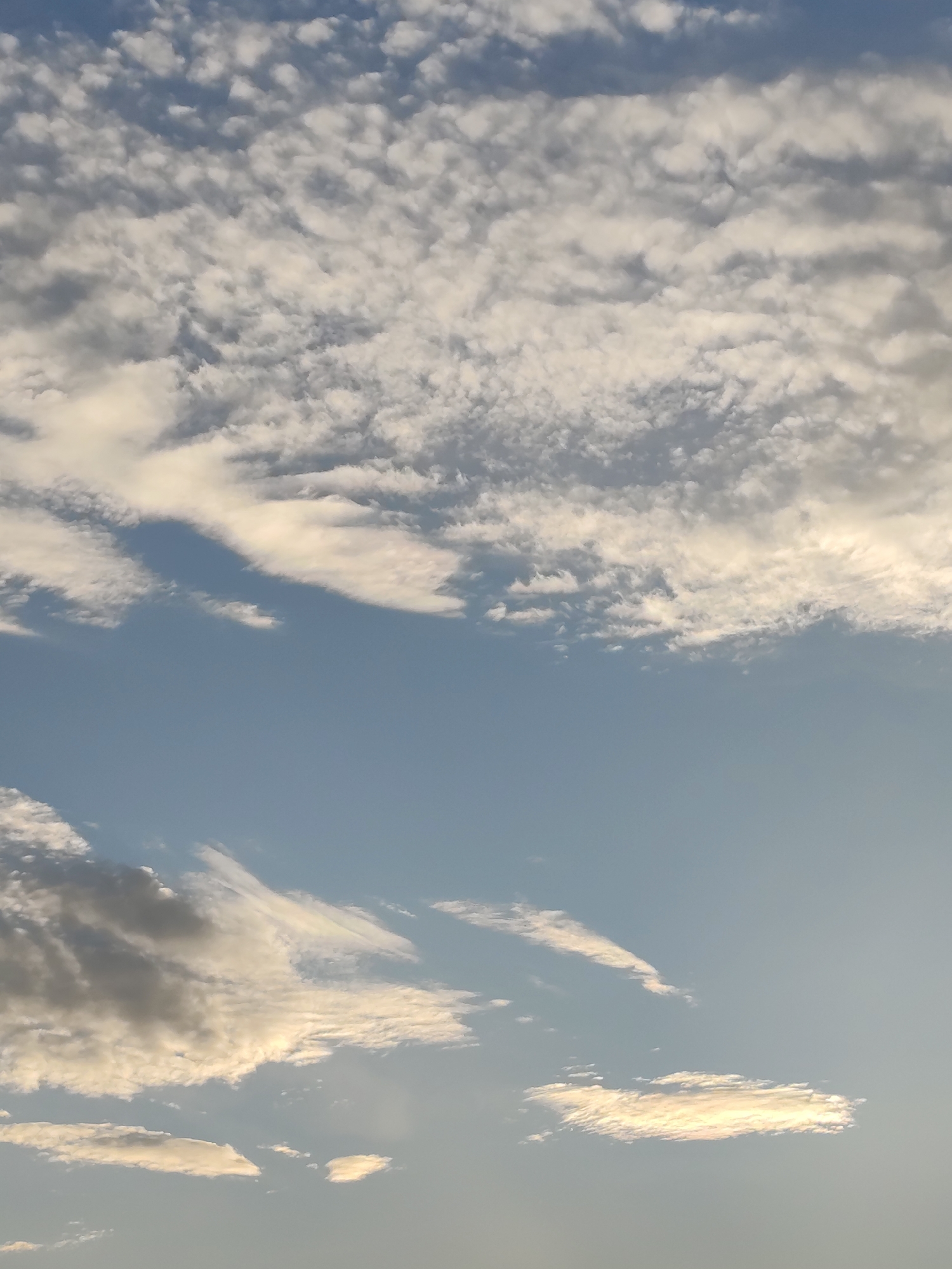The image shows a sky filled with scattered clouds under a clear blue sky. The clouds vary in density and shape, with some appearing wispy and others more substantial. The lighting suggests it might be late afternoon or early morning, as some clouds are tinged with yellow and white from the sunlight. There are no people, subjects, or objects visible in the image, only the sky and its atmospheric conditions. The scene is a natural outdoor environment.