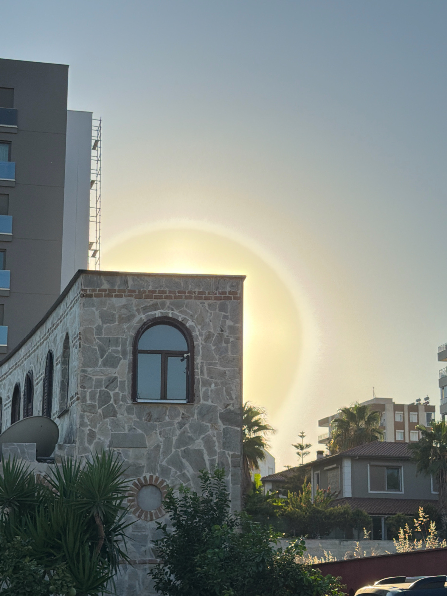 The image shows a sun halo phenomenon occurring in the sky above a cityscape. The halo, a bright ring of light around the sun, is visible in the upper portion of the image. In the foreground, a building with a stone facade and arched windows is prominent. To the left of this building, a taller, modern gray building with balconies is partially visible. The background features residential buildings and palm trees, suggesting a warm climate. The overall impression is of an urban environment bathed in the soft light of either sunrise or sunset, with the sun halo adding a natural spectacle to the scene.