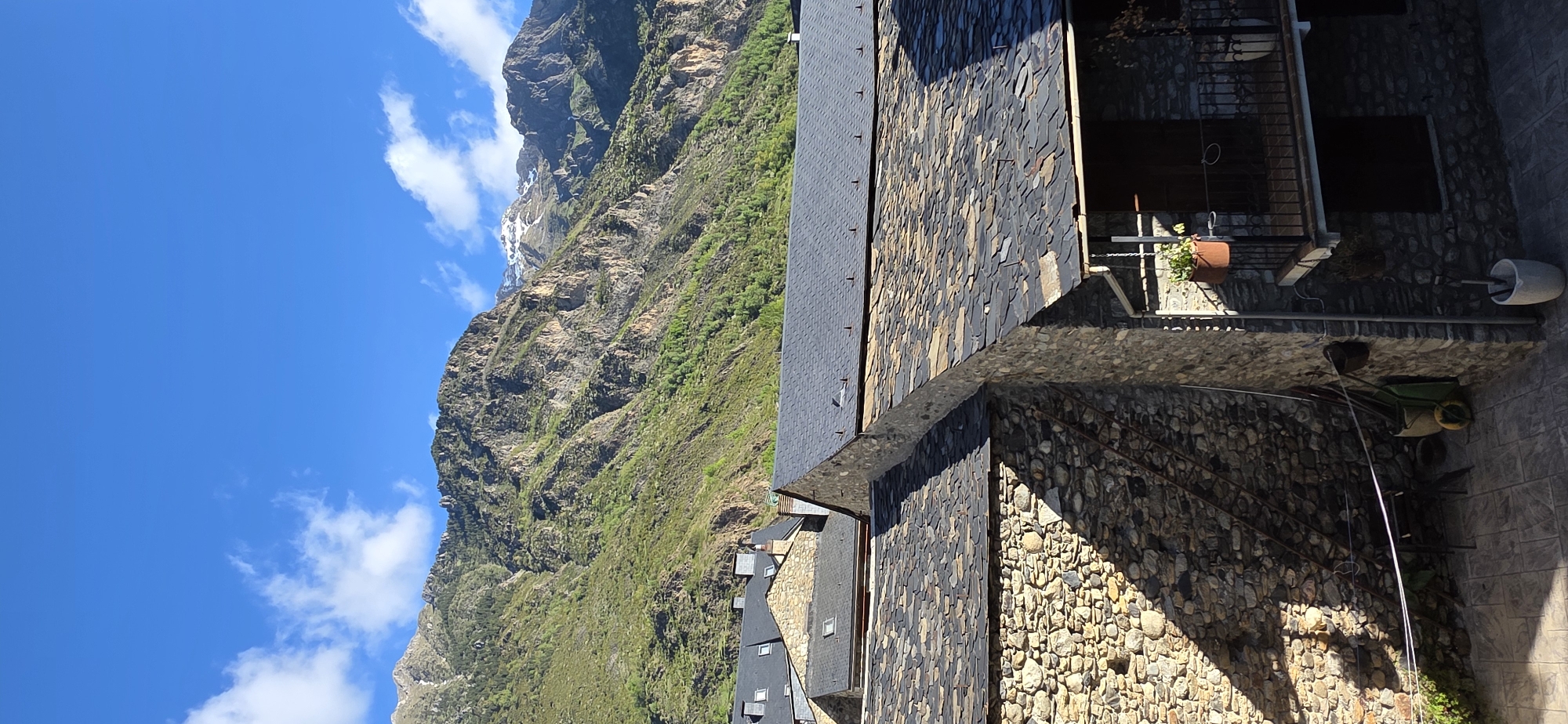 The image shows a mountain landscape juxtaposed with residential architecture, potentially located in the city of Benasque, Spain. The mountains are rugged, with patches of green vegetation and some snow near the peak. The sky is a clear blue with scattered clouds. Adjacent to the mountain range, a building is visible. It is constructed with a stone facade and a slate roof. The building appears to be a residential structure, as evidenced by a balcony with potted plants and a stone patio containing gardening tools. The overall setting combines natural mountainous terrain with village architecture.