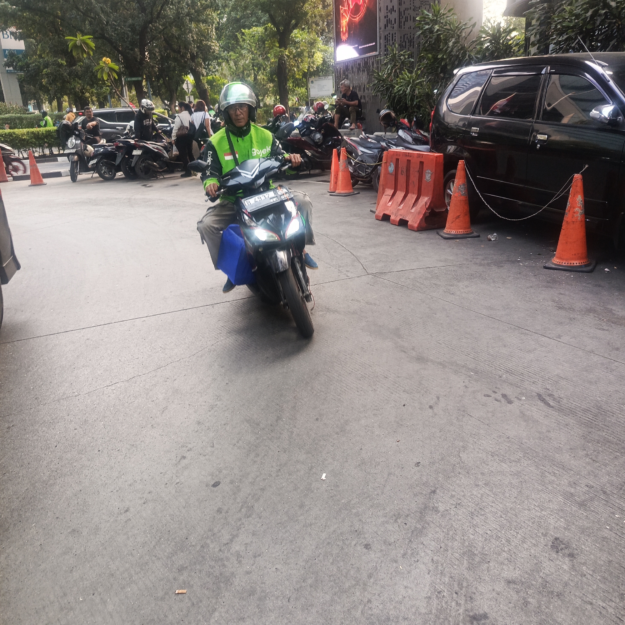 The image depicts a street scene in South Jakarta, Indonesia, with a focus on a motorcycle courier. The courier, wearing a green jacket with "gojek" branding and a white helmet, is seated on a black scooter, navigating the paved road. The scooter has a license plate reading "B 4937". A blue bag is strapped to the back of the scooter. In the background, several other motorcycles are parked, and a black MPV is visible. Orange traffic cones and concrete barriers are placed along the side of the road, indicating a potential construction or traffic diversion area. Trees and foliage border the street, and a building with a large illuminated screen is partially visible in the upper right quadrant. The overall environment appears to be an urban setting, possibly a commercial or high-traffic area.