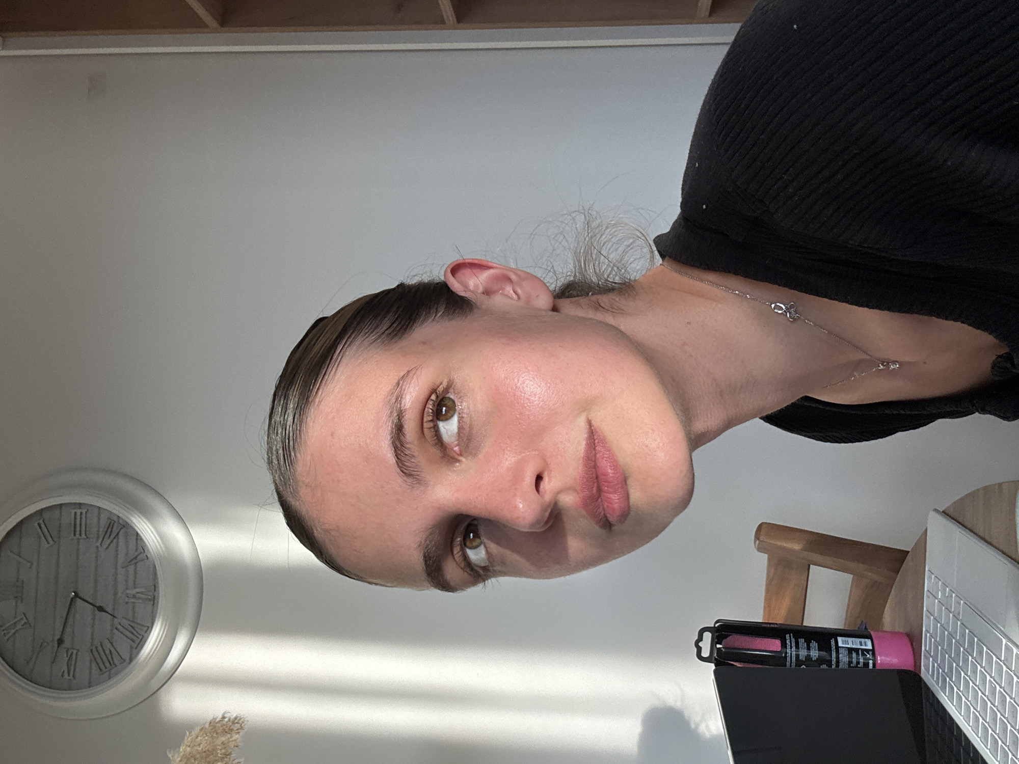 A woman with brown eyes and slicked-back hair is the primary subject. She is wearing a black shirt and a delicate silver necklace. In the background, a clock with Roman numerals reads approximately 7:26. A laptop, a pink highlighter, and a wooden chair are partially visible on a desk. The setting appears to be indoors against a light gray wall. The location is Unknown, Unknown.