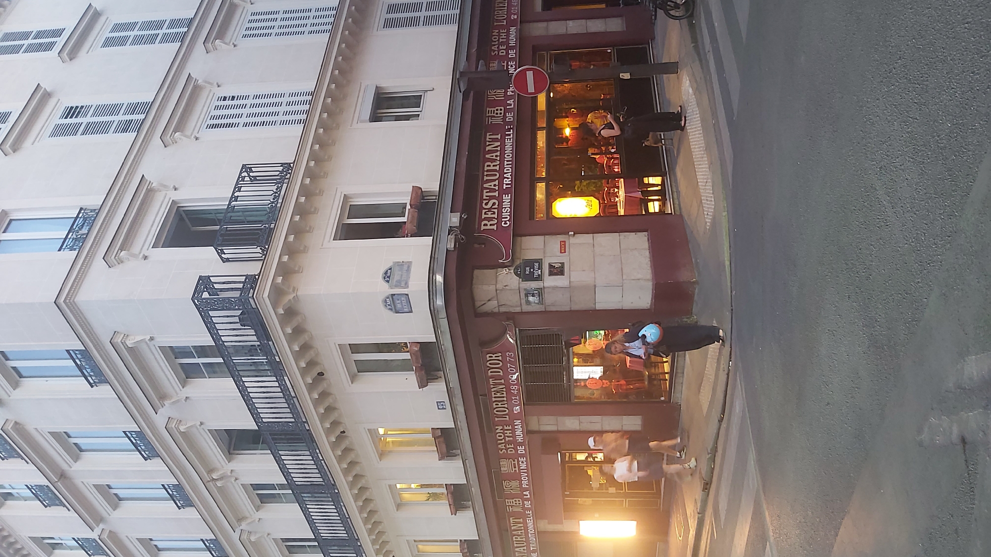 The image shows a street corner in Paris, France, featuring the corner of a building with a restaurant on the ground floor. The building is light-colored stone with multiple stories, exhibiting classic Parisian architecture with balconies and shutters on the upper floors. The restaurant's facade is primarily dark red with prominent signage. On the left side, the sign reads "RESTAURANT 貓 貓 貓 SALON DE THE LORIENT DOR 01 48 09 07 73 CUISINE TRADITIONNELLE DE LA PROVINCE DE HUNAN". On the right side, the sign reads "RESTAURANT 貓 貓 貓 SALON DE THE". Visible street signs indicate "RUE RICHER" and "RUE TREVISE".
Several people are present. On the left, two individuals are walking, appearing to be patrons or passersby. One is wearing a light-colored t-shirt and shorts, and the other is wearing a white cap and dark clothing. Near the entrance, a woman is standing and looking at her phone, holding a blue helmet. Further inside the restaurant, other patrons can be seen at tables. A small dog is standing on the sidewalk near the entrance on the right side. A traffic light with a red circle and a white horizontal bar, indicating no entry, is mounted on a pole on the right side of the image, near the street. A bicycle or motorcycle is parked on the far right edge of the frame. The street in the foreground is paved with asphalt and includes a pedestrian crossing. The lighting suggests it is late afternoon or early evening.