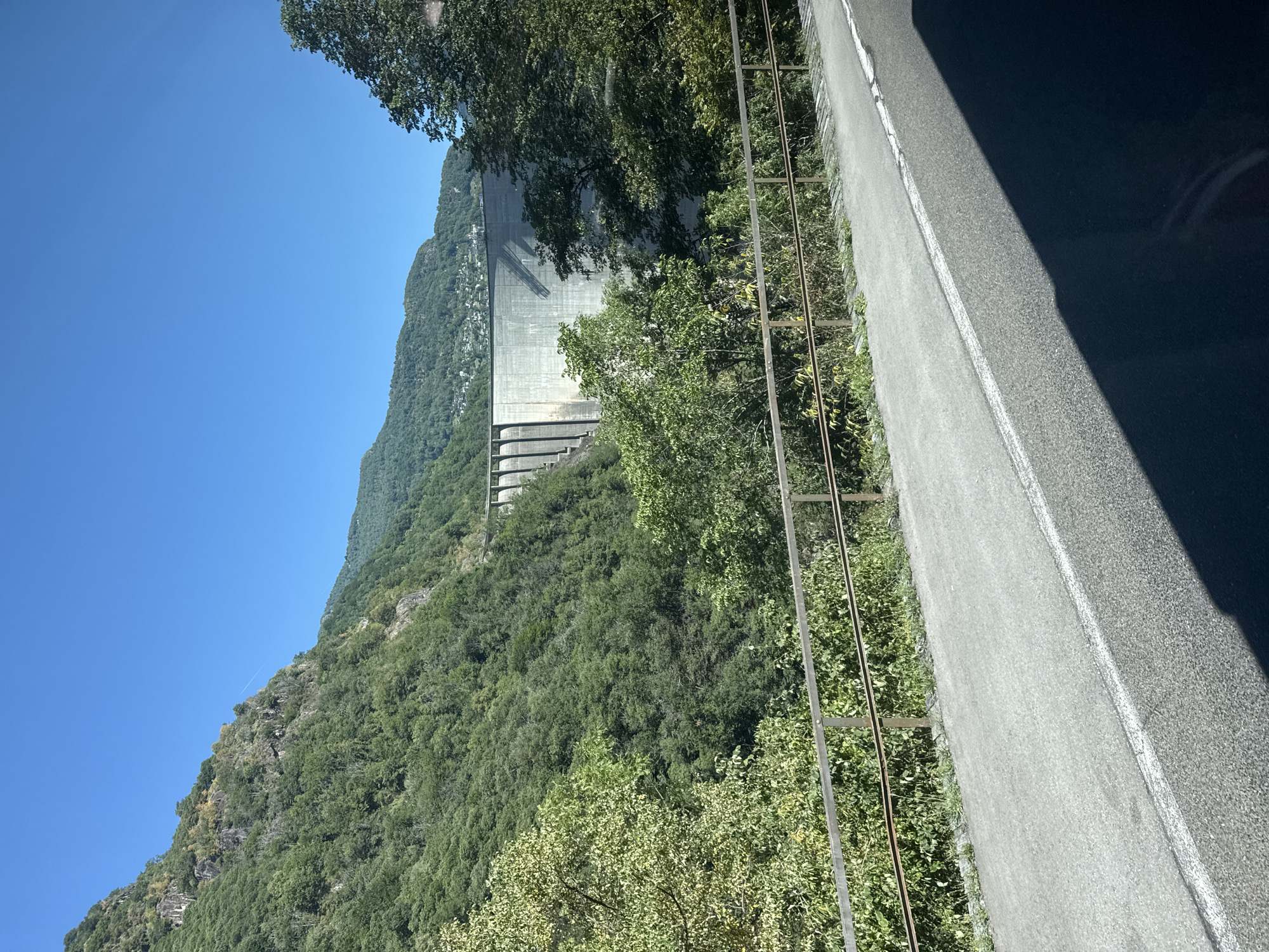 The image shows a large concrete dam in a mountainous, forested area. The dam is curved and has a series of vertical structural elements visible. The surrounding landscape is covered in lush green trees, with steep rocky slopes on the left side. The sky is clear and blue. In the foreground, there is a paved road with a white line marking its edge, and a metal railing separates the road from the vegetated area below. A shadow of an object, possibly a vehicle, is cast across the road. The location is identified as the city of Gordemo, Switzerland.