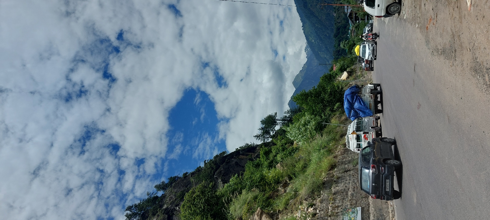 This is a photograph taken in Kalath, India, depicting a mountainous landscape with a road and vehicles. The sky is filled with white, puffy clouds against a bright blue backdrop. On the left side of the image, a steep, rocky mountain covered in lush green trees and vegetation dominates the scene. A paved road runs horizontally across the foreground, with several vehicles on it.
In the foreground, a dark grey car is visible, facing away from the camera. Further down the road, a white minibus is parked, and behind it, a white truck with a large blue tarp covering its load is also stopped. Several other cars and two motorcycles are seen further ahead on the road, with one motorcycle carrying a rider. The road is lined with trees and some buildings are visible in the distance on the right side of the frame, nestled within the valley. The overall environment suggests a scenic highway route through a hilly region.
