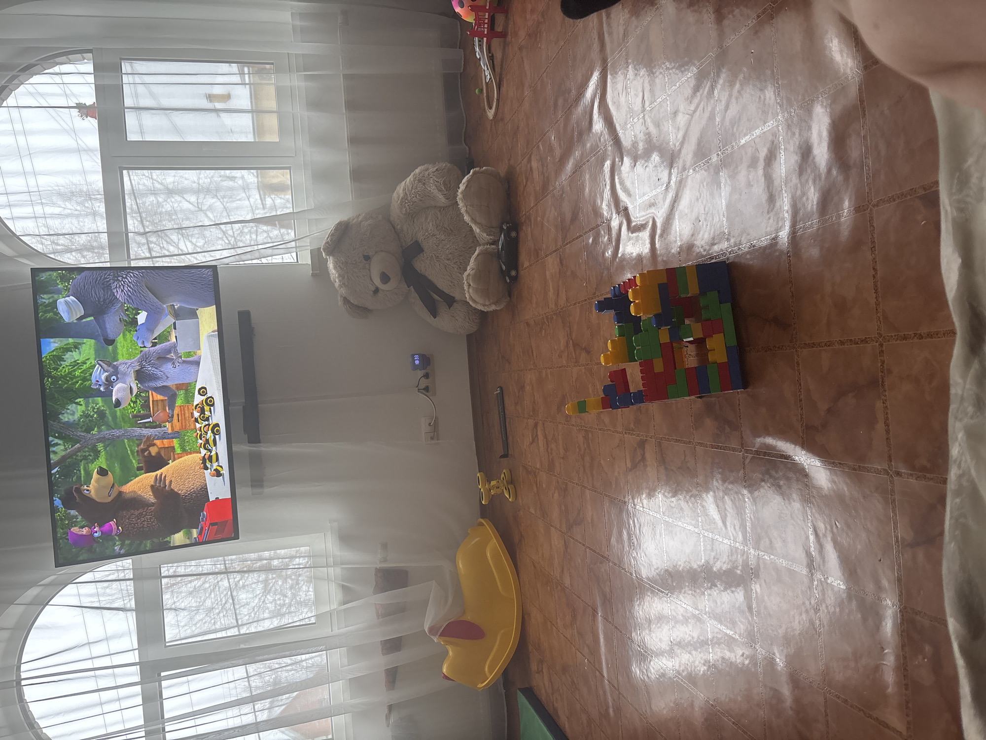 The image depicts an indoor scene in what appears to be a child's play area or living room, located in Ферма, Russia. The room features a tiled floor with a brown, possibly terracotta, color and a glossy finish reflecting some light.
Key subjects include a large, light brown stuffed teddy bear with a black bow tie, sitting upright on the floor near a wall. In front of the bear, a television is displaying an animated children's show, clearly identifiable as "Masha and the Bear," featuring characters like Masha, the Bear, and a wolf. To the right of the teddy bear, a colorful structure made from various red, blue, green, and yellow building blocks is constructed on the floor. Further into the room, a bright yellow plastic rocking toy, possibly a duck or swan, is visible, along with a small yellow wheeled toy nearby. A small black toy car is also near the teddy bear.
The room is illuminated by natural light entering through large windows covered by sheer white curtains. The windows have white frames, with one section featuring a distinctive semicircular top. Outside, bare tree branches suggest a season like late autumn or winter. A small, wall-mounted digital clock with blue numerals reads "18:33," indicating the early evening. No people are visible in the scene, suggesting a quiet moment in a home. The overall atmosphere is neat and organized, with toys spread out, implying recent or upcoming play.