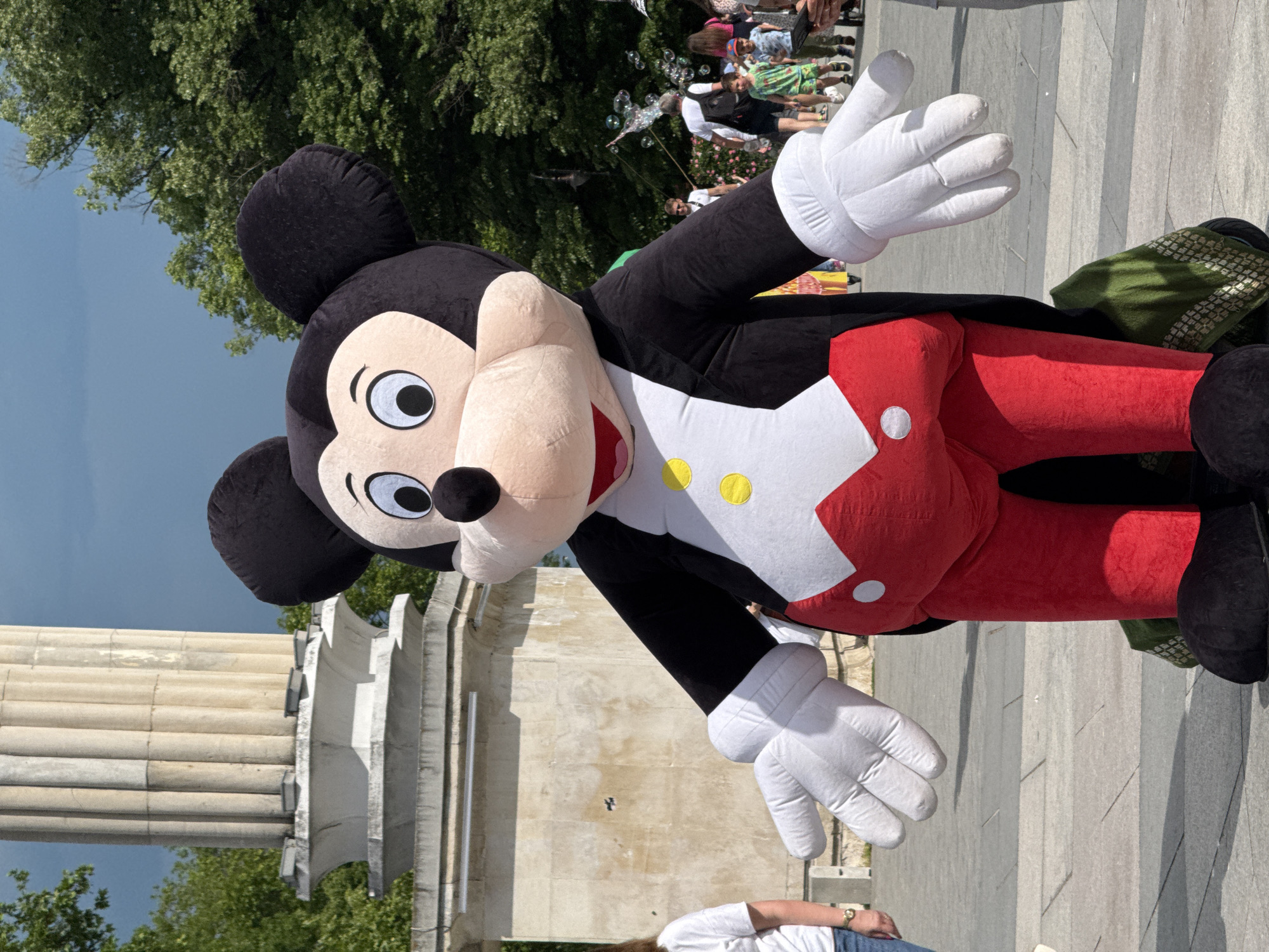 A vertical, full shot shows a large Mickey Mouse mascot in front of a monument in Varna, Bulgaria. Mickey is wearing a black coat with white lapels, a white shirt with yellow buttons, and red pants. His arms are outstretched in a welcoming gesture. The statue behind him is white with a stone facade and some trees visible in the background. The background also features people and soap bubbles. The ground is paved with stone. The image appears to be taken at ground level.