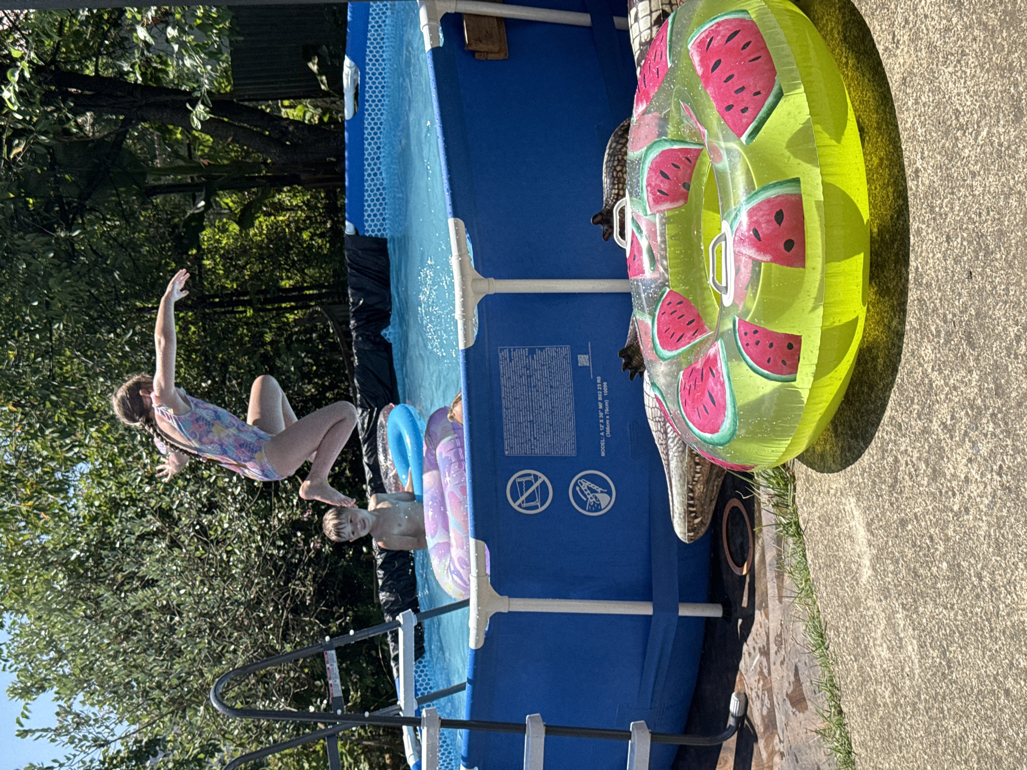This media file captures a lively outdoor scene featuring children enjoying a summer day by an inflatable pool. A young girl in a colorful swimsuit is captured mid-air, mid-jump into the blue pool, her arms outstretched. Beside her, a young boy, topless and with a surprised expression, is partially submerged in the water, leaning on inflatable rings.
The pool itself is blue with white supports, and a prominent inflatable ring with a watermelon pattern is visible, leaning against the pool's side. A realistic-looking inflatable alligator is also partially visible, adding a playful element to the scene. The surrounding area is lush with green trees and foliage, suggesting a backyard setting. The ground around the pool is a concrete patio, with some grass growing at the edge.
The lighting suggests it's a sunny day, casting shadows from the trees and the pool. The overall mood is one of fun and recreation. Text visible on the pool's side indicates "MODEL: A 12" X 30 MF B02 25 RO (366cm x 76cm) 10096". The scene implies a warm, bright day, perfect for swimming and playing in an inflatable pool.