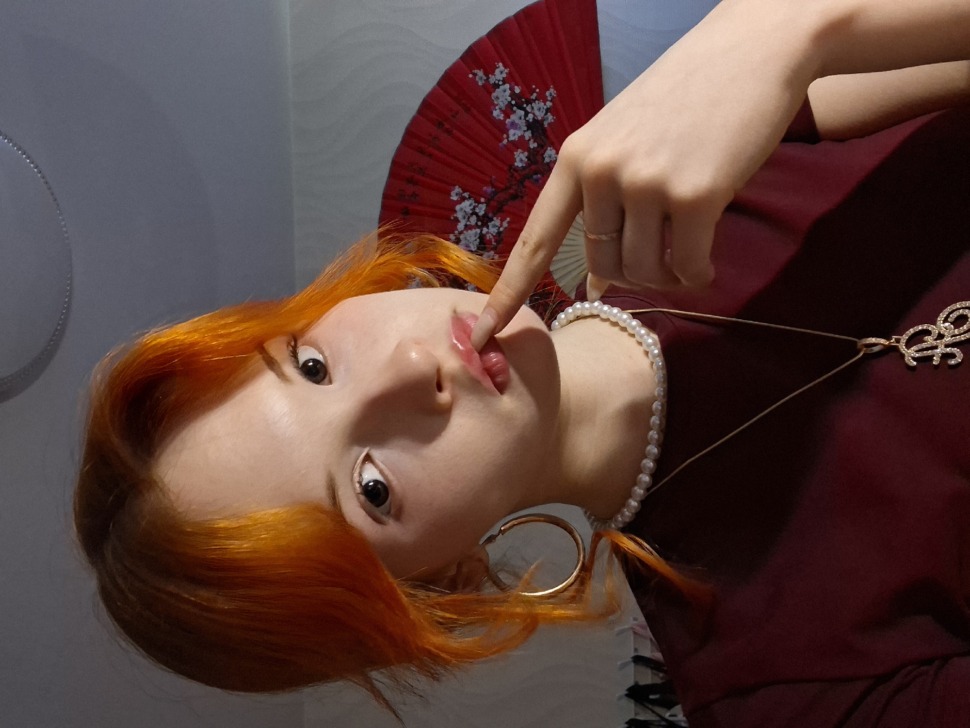 A woman with reddish-orange hair is pictured indoors, with her head tilted to the left. She is wearing a dark maroon shirt, a pearl necklace, a gold necklace with a pendant, and gold hoop earrings. Her right index finger is touching her lip. A red fan with floral patterns is visible behind her. The background includes a gray wall, a white surface, and a partially visible black object. The location is Chaikovsky, Russia.