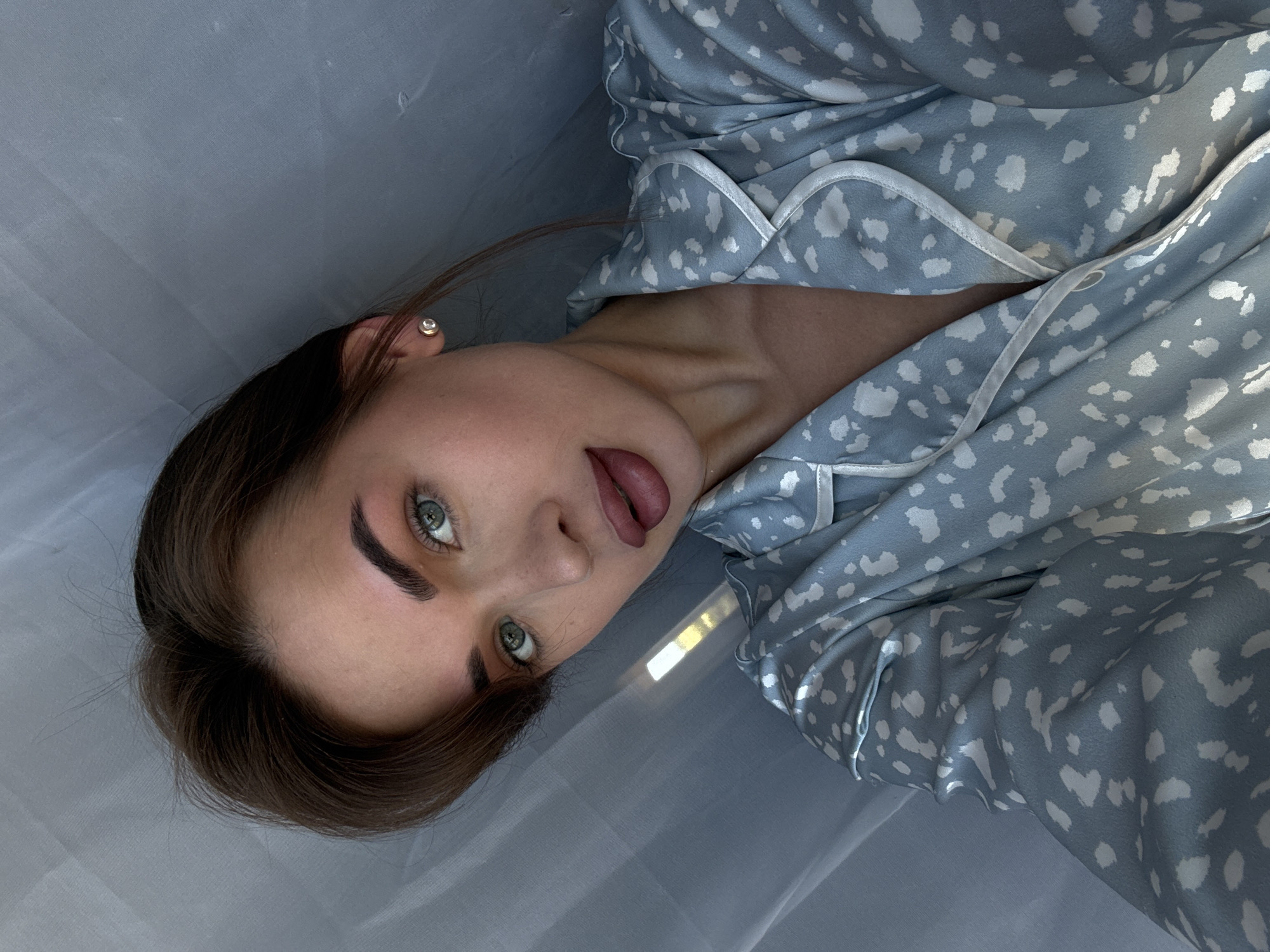 A young woman with brown hair and green eyes is lying on a light gray surface, possibly a bedsheet. She is wearing a light blue pajama top with white spots and white piping. Her head is tilted to the right, and she is looking directly at the camera with her mouth slightly open, revealing her tongue. A single earring is visible in her left ear. A patch of bright sunlight is visible on the gray surface. The location is unknown, and the suggestion of Лобаново, Russia is unsupported by the image.