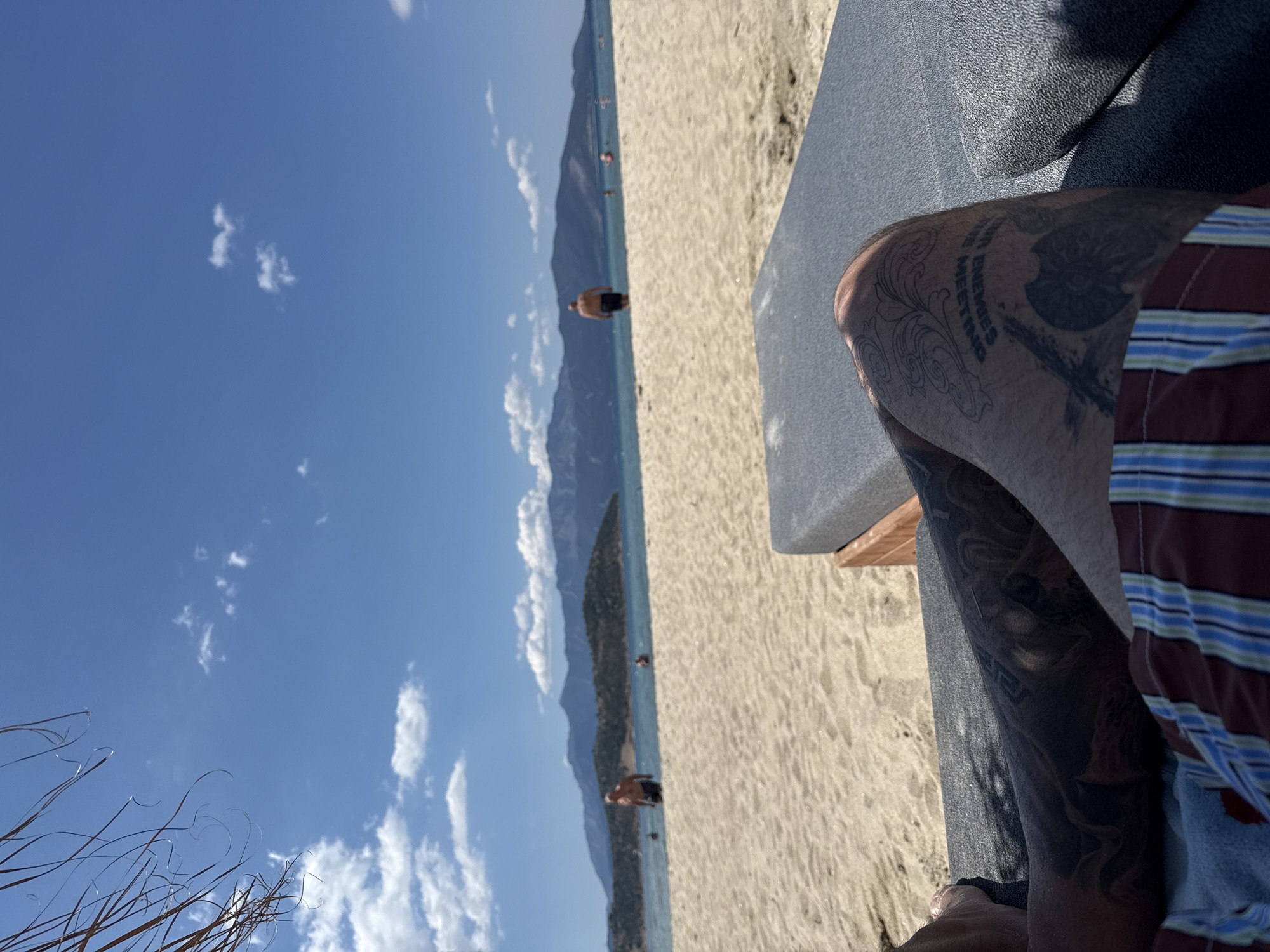 The image depicts a beach scene in Keramoti, Greece. In the foreground, a person's tattooed legs and striped shorts are visible, resting on a lounge chair. The beach is composed of light-colored sand, with a lounge chair situated on it. In the background, the blue sea stretches towards a distant coastline featuring a range of mountains. Several individuals are visible in the water and on the beach, some wading in the sea and others walking. The sky is a clear blue, dotted with scattered white clouds. The overall setting suggests a sunny day at a coastal resort.