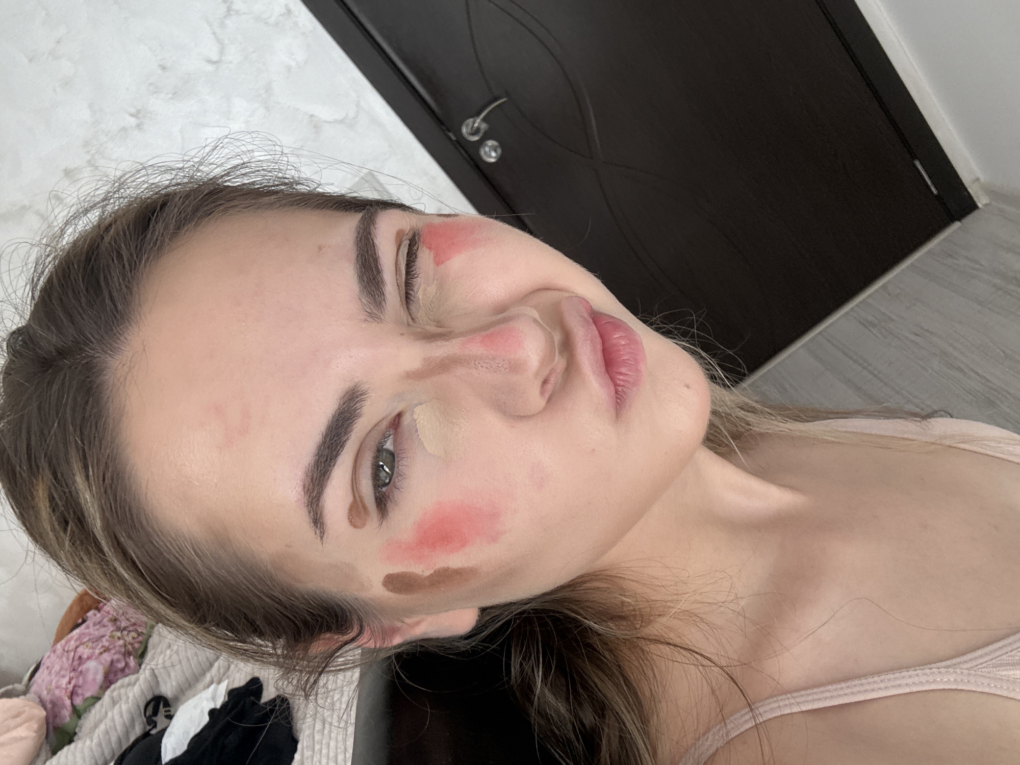 A young woman is pictured indoors, likely in a private residence. She has fair skin, brown hair, and green eyes, and is wearing a pink tank top. Her face has several blotches of makeup primer, blush, and contour cream. The background includes a white wall and a dark brown door. The image does not show a location in Blagoevgrad, Bulgaria.