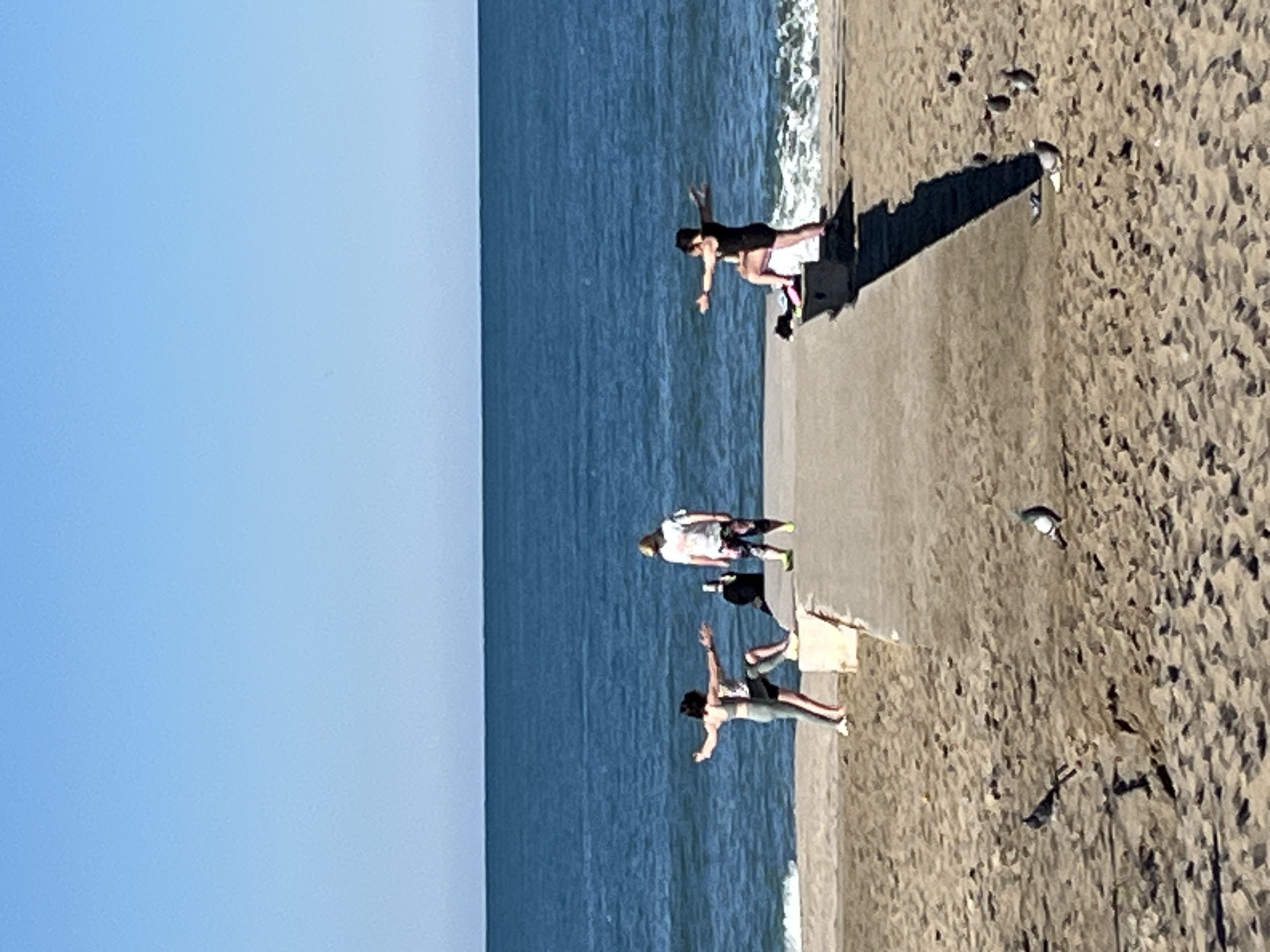 The image depicts a beach scene in Alanya, Turkey. The scene shows three individuals on a concrete embankment parallel to the water's edge. Two of the individuals have their arms outstretched, possibly posing. One individual in the middle of the frame is dressed in a light colored t-shirt with dark shorts. The beach consists of wet sand, interspersed with small puddles and some seagulls. The sea is light blue, with a cloudless blue sky occupying the upper left of the image.