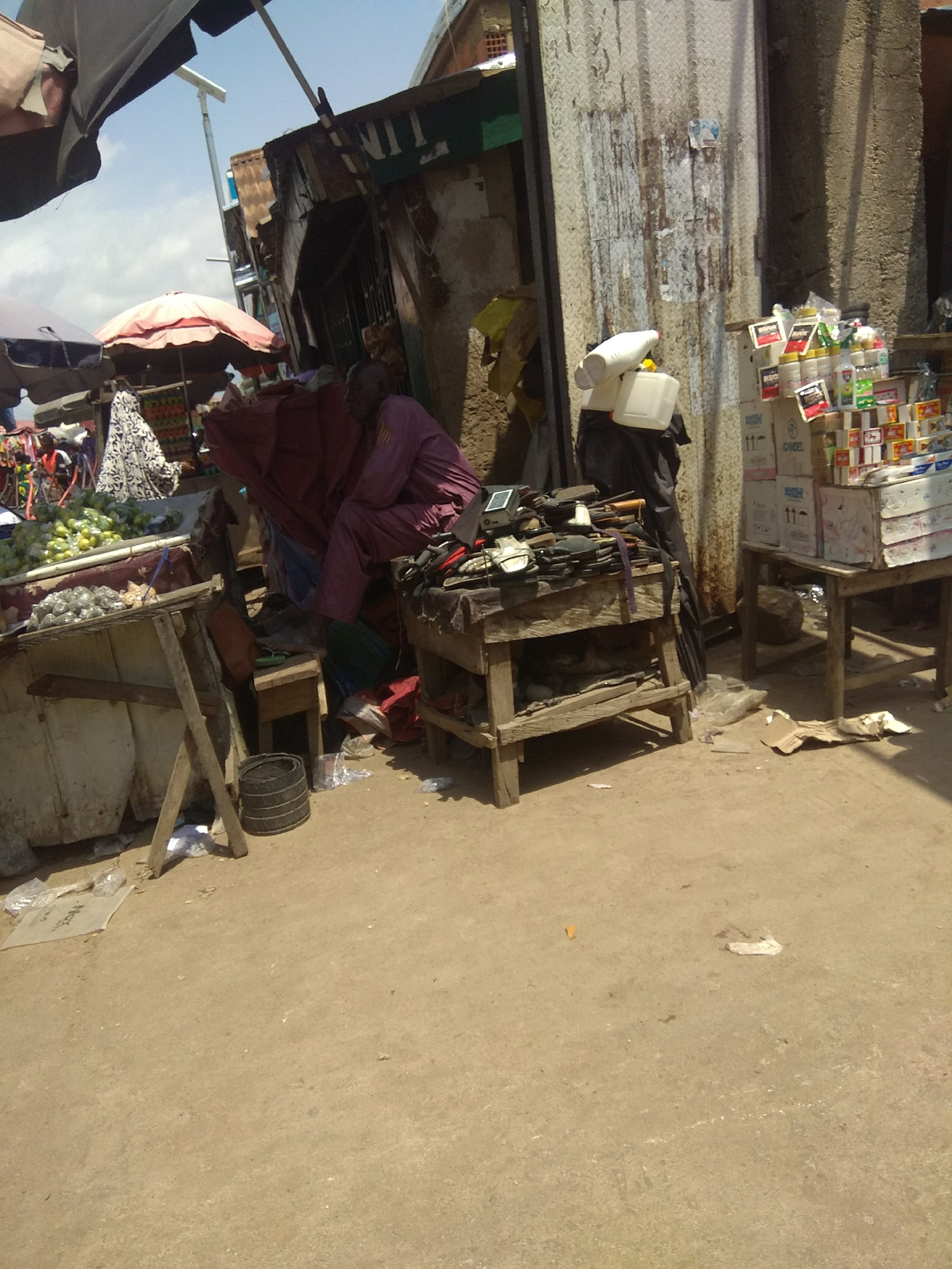 A medium shot captures an outdoor market scene, likely in Jalingo, Nigeria. The sun is shining, indicating daytime. The foreground is a dusty, light brown ground. To the left, a worn wooden stall displays various goods, including what appears to be a pile of produce, possibly limes or small green apples, and some metal objects. Behind this stall, a person in a patterned white and black garment is partially visible, as is a pink umbrella.
In the center of the image, a person in a mauve or purple traditional outfit is seated. They are facing away from the camera and towards a wooden table laden with what look like tools, possibly for shoe repair, and other small items. This table has a rustic, weathered appearance. To the right of the seated person, a dark fabric is draped, and further right, a tall structure with corrugated metal siding stands. This structure is weathered and has some peeling paint or posters.
In front of the corrugated structure, several boxes are stacked, some containing what appear to be bottled goods. A white plastic container and a white, nozzle-like object are also visible near the boxes. The overall scene suggests a bustling marketplace with various vendors and their wares. The presence of umbrellas, stalls, and stacked goods points to commercial activity. The ground is strewn with some debris, including plastic bags and paper, common in such environments. The lighting and shadows suggest a bright, sunny day. No specific text is clearly legible.